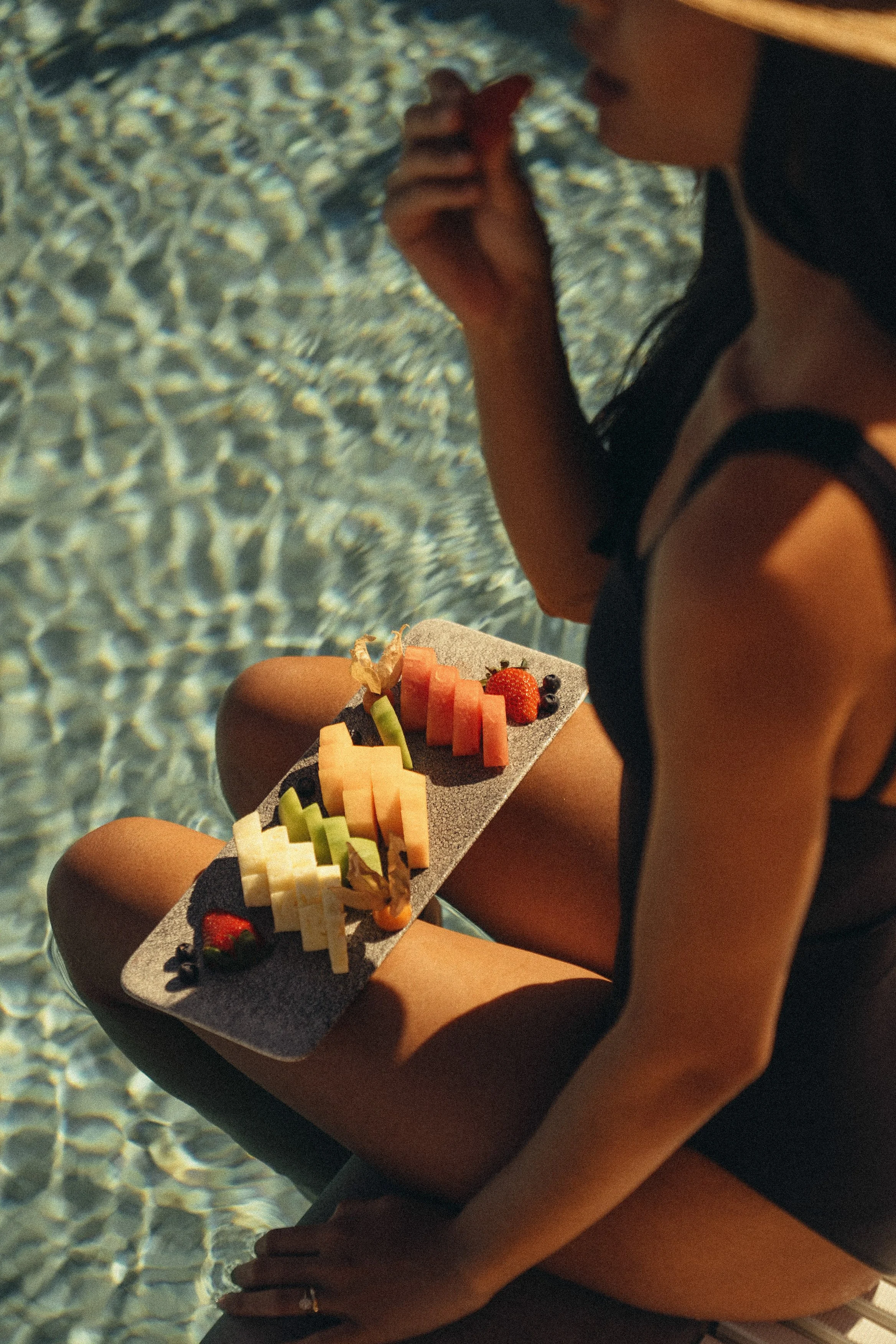 A woman in a black swimsuit sitting near water, holding a plate of assorted fruit slices including watermelon, cantaloupe, honeydew, strawberries, blueberries, and pineapple.