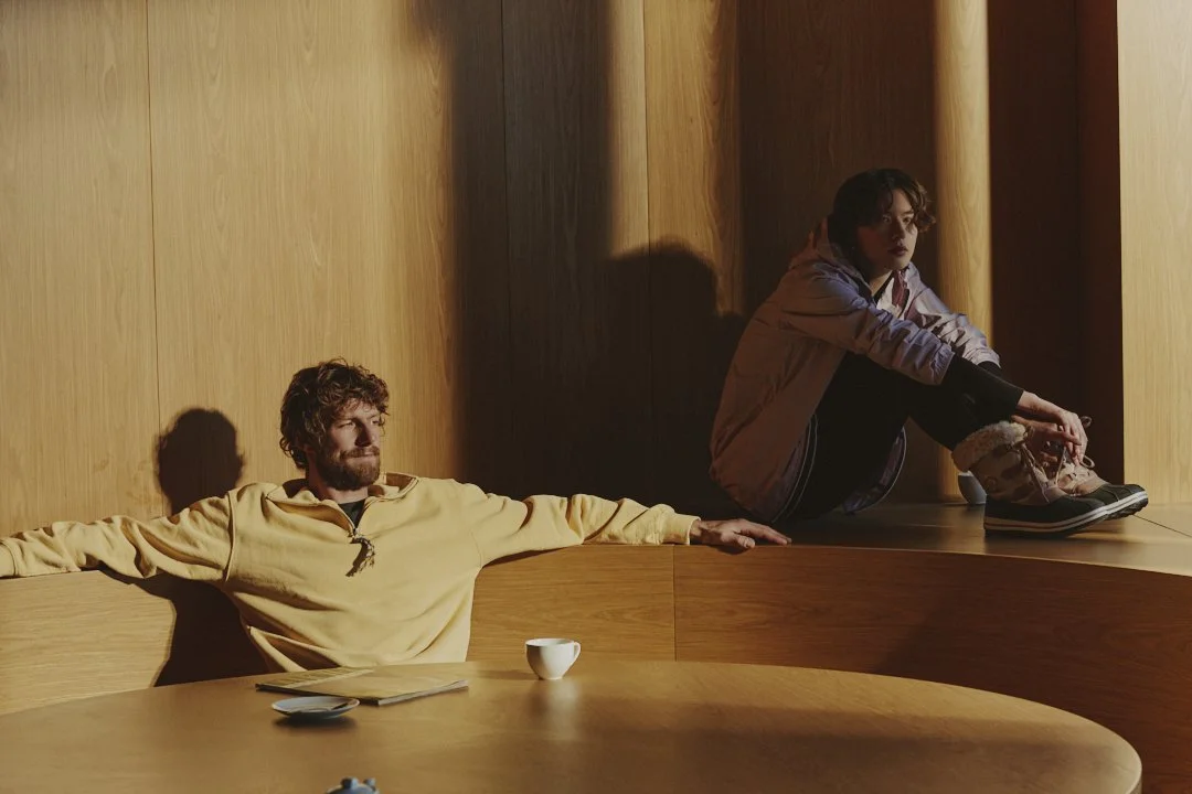 Two people sit in a room with wooden walls. One man in a yellow shirt is leaning back against the wall with eyes closed, and a woman in a purple jacket sits on the floor nearby, looking away with a serious expression.