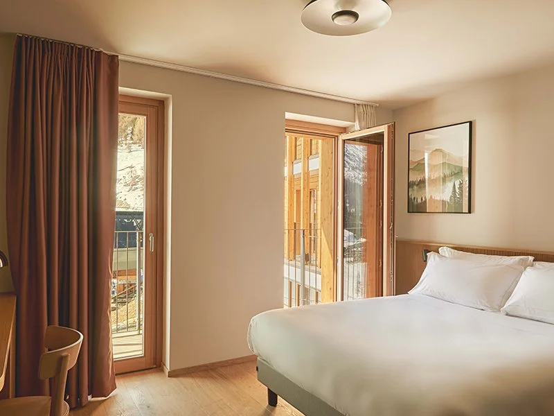 A hotel room with a large bed, wooden headboard, white bedding, and pillows. There are two windows: one with closed brown curtains and another open door leading to a balcony with wooden railings and a view of a building under construction.