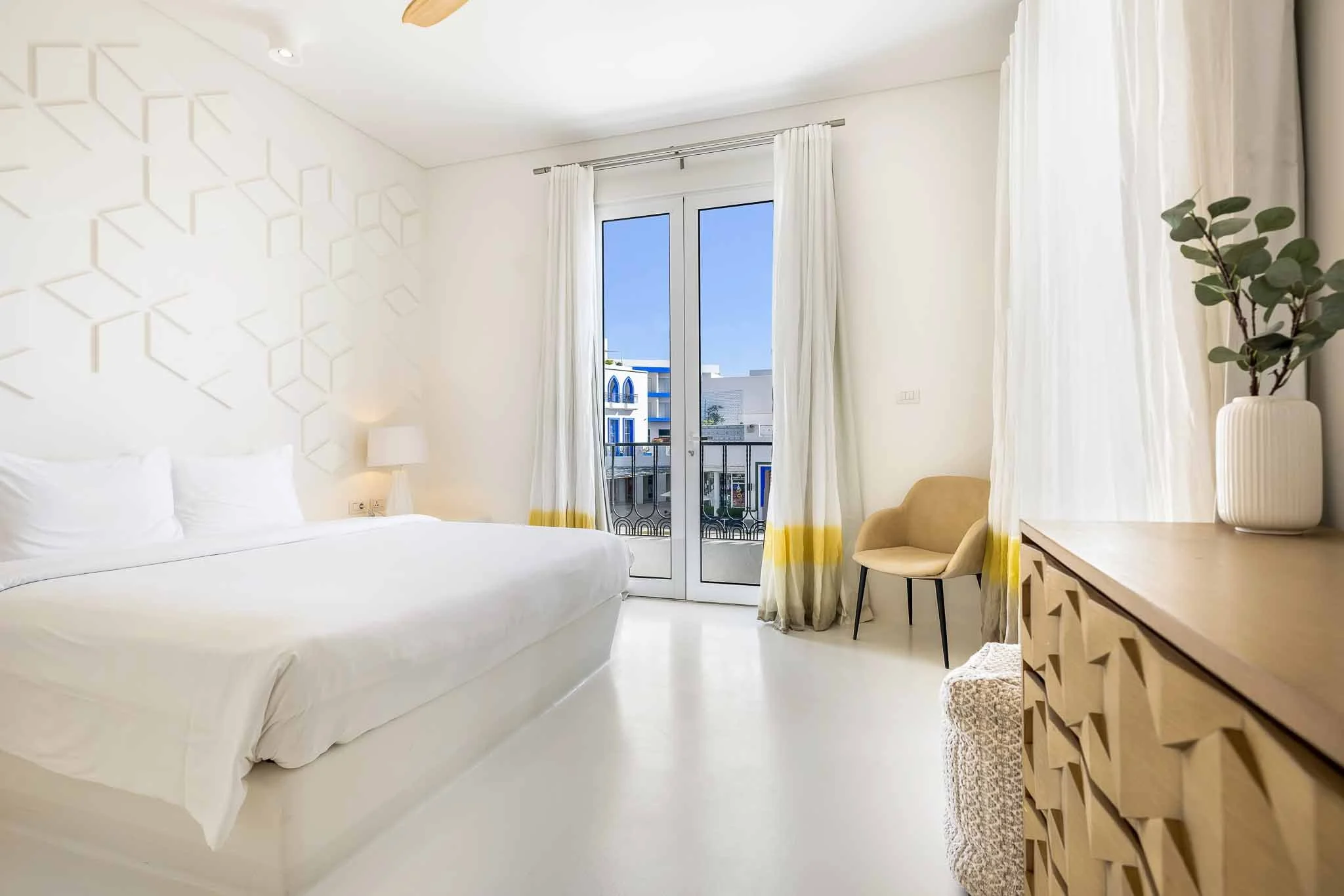 A bright hotel room with white walls and bedding, a large window with white curtains, a beige chair, a wooden desk with a decorative pattern, a potted plant, and a balcony view of white buildings with blue accents.