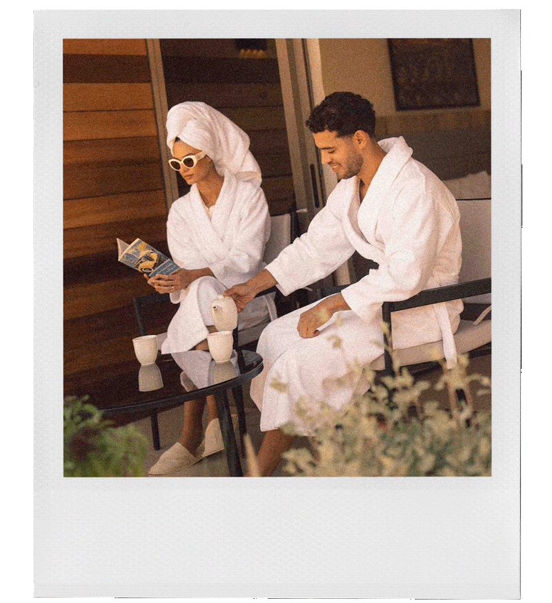 Two people in bathrobes sitting at a glass table on a patio, one reading a magazine and the other pouring a drink, in a relaxing outdoor setting.