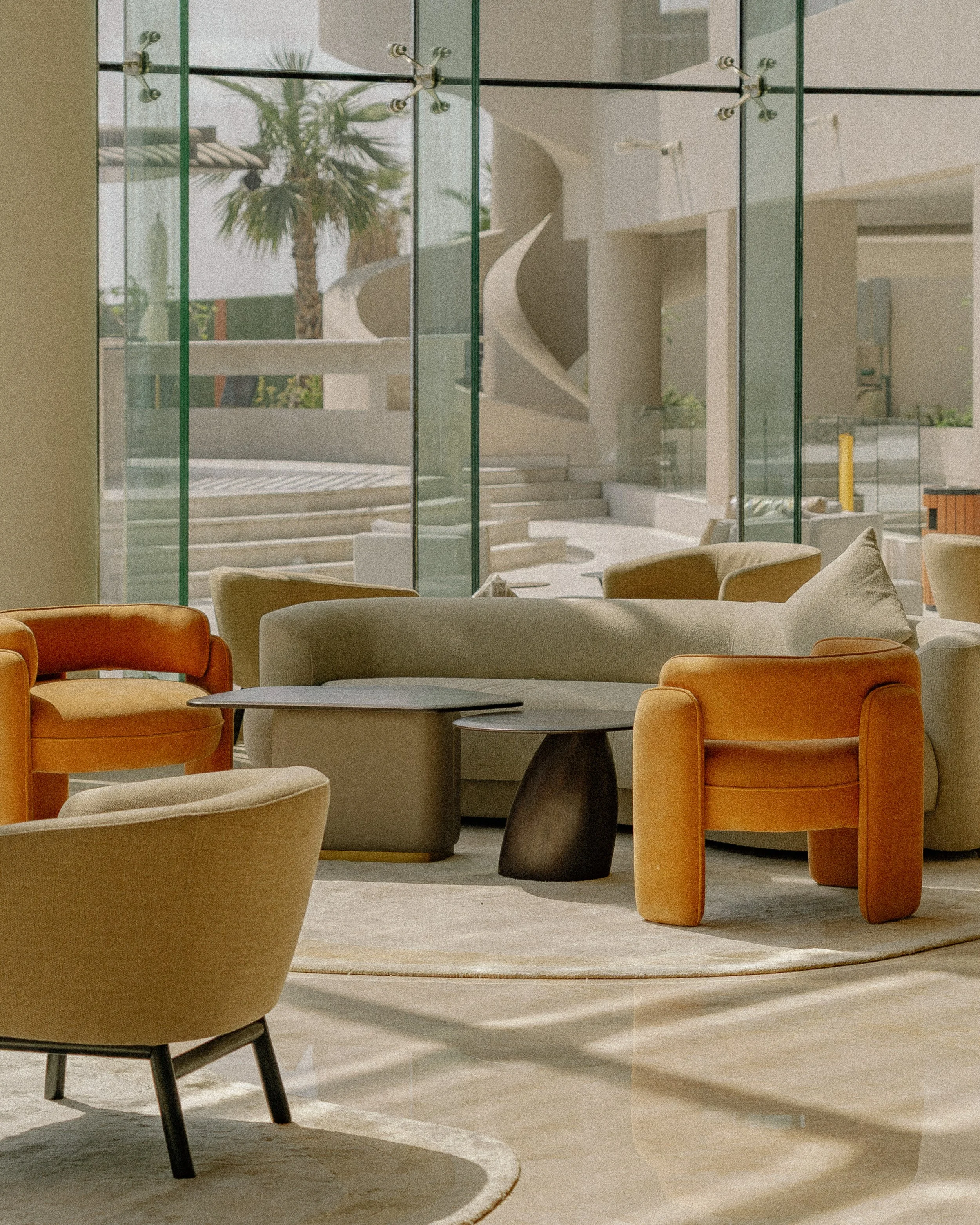 Living room with beige armchairs, orange chairs, a round coffee table, large glass windows showing outdoor palm trees, and a spiral staircase.