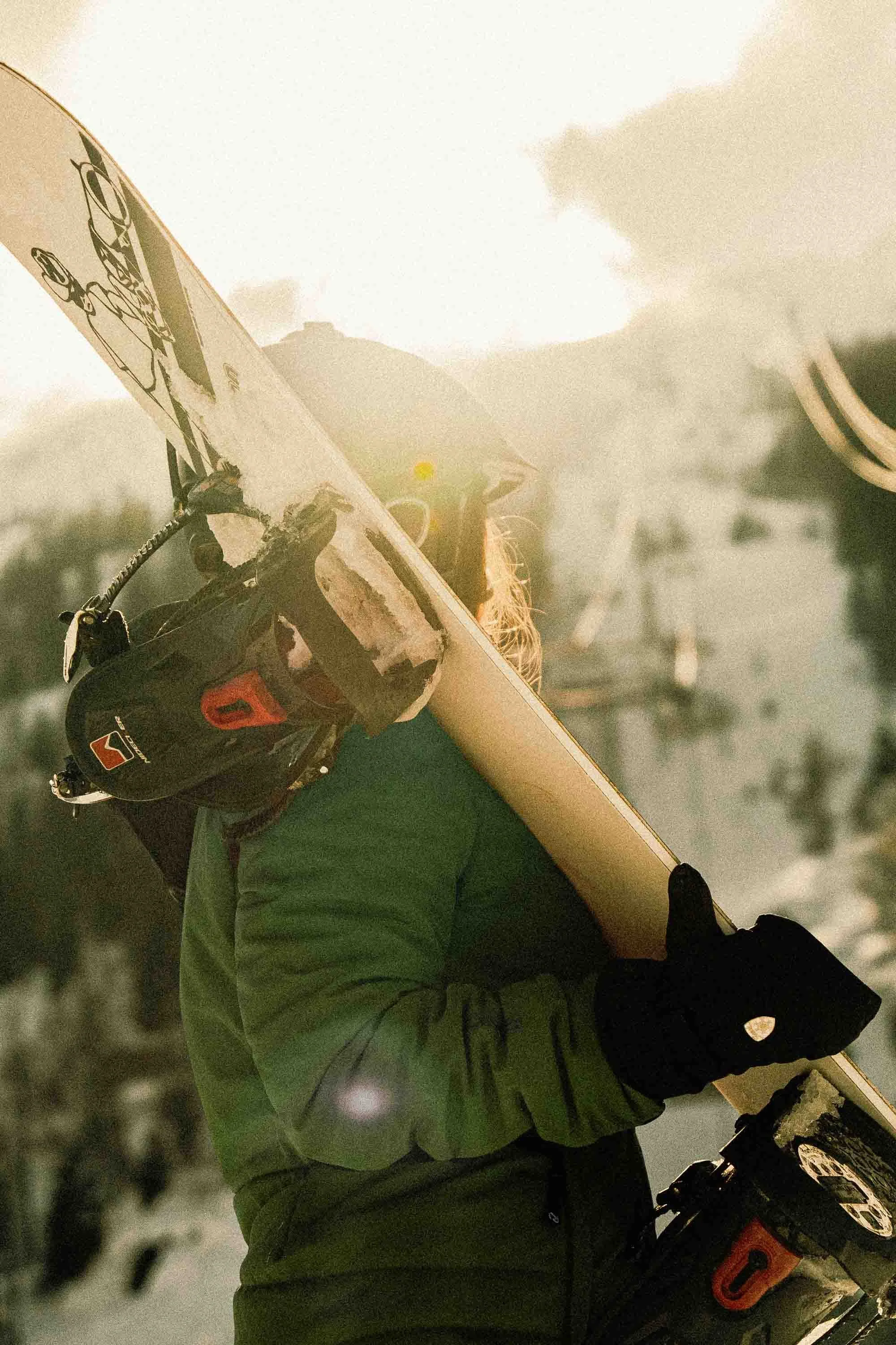 Person wearing a green jacket and black gloves holding a yellow surfboard, with clouds and trees in the background.