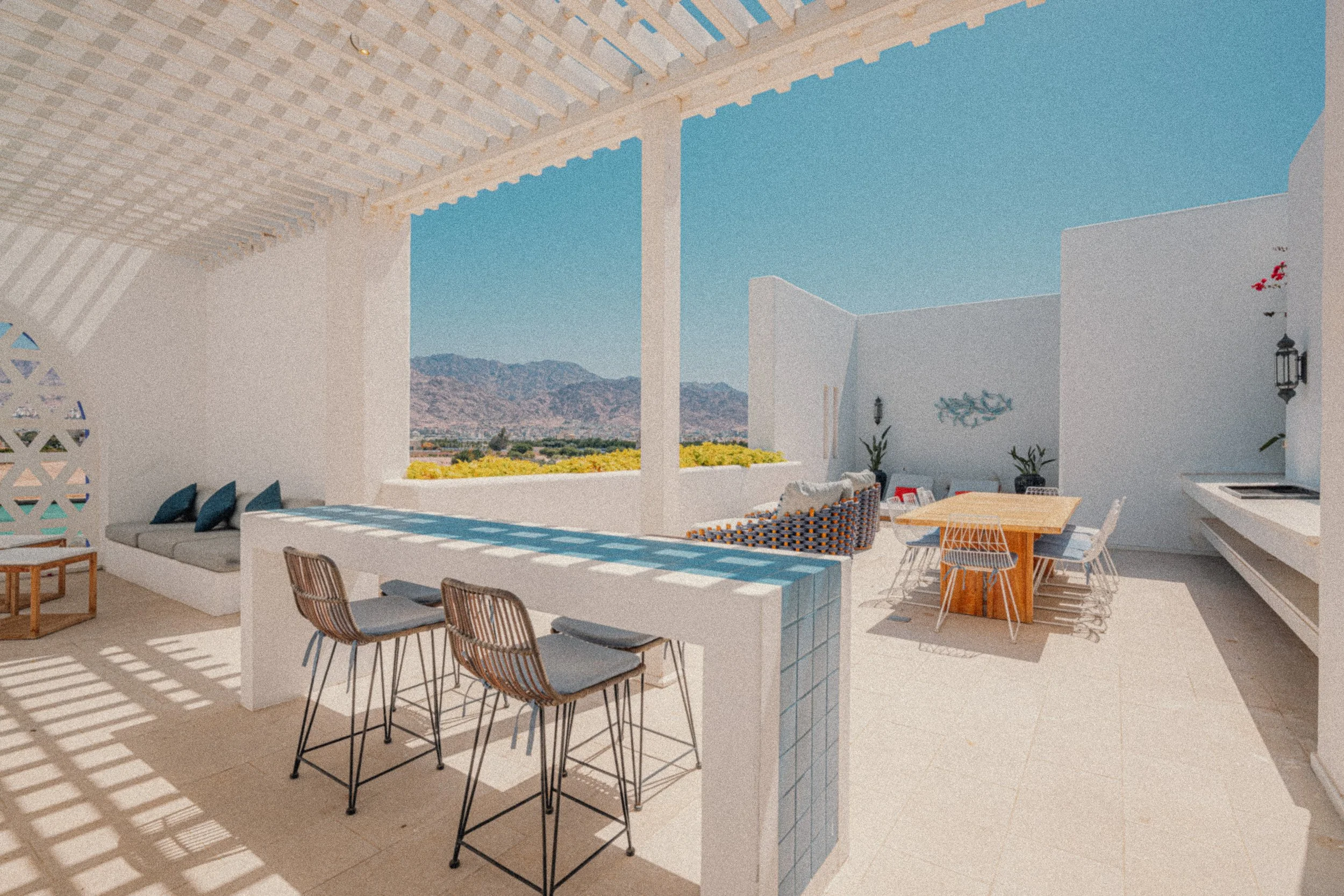 Outdoor rooftop terrace with seating area, tables, and mountain view in the background, shaded by a lattice pergola.