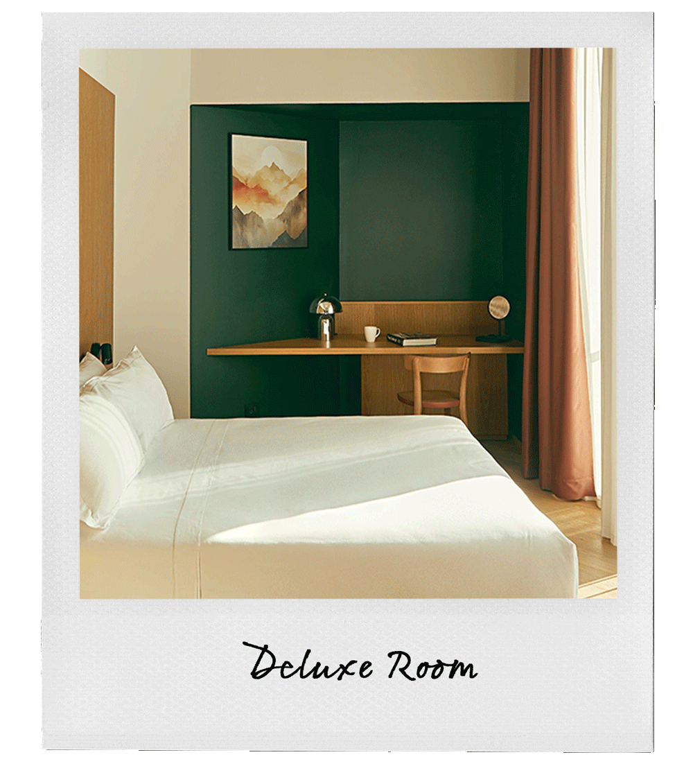 A hotel room with a neatly made bed and a built-in wooden desk against a dark green accent wall, featuring a framed mountain landscape painting, a modern desk lamp, a white mug, a closed book, and a small decorative globe, illuminated by natural light from a window with beige curtains.