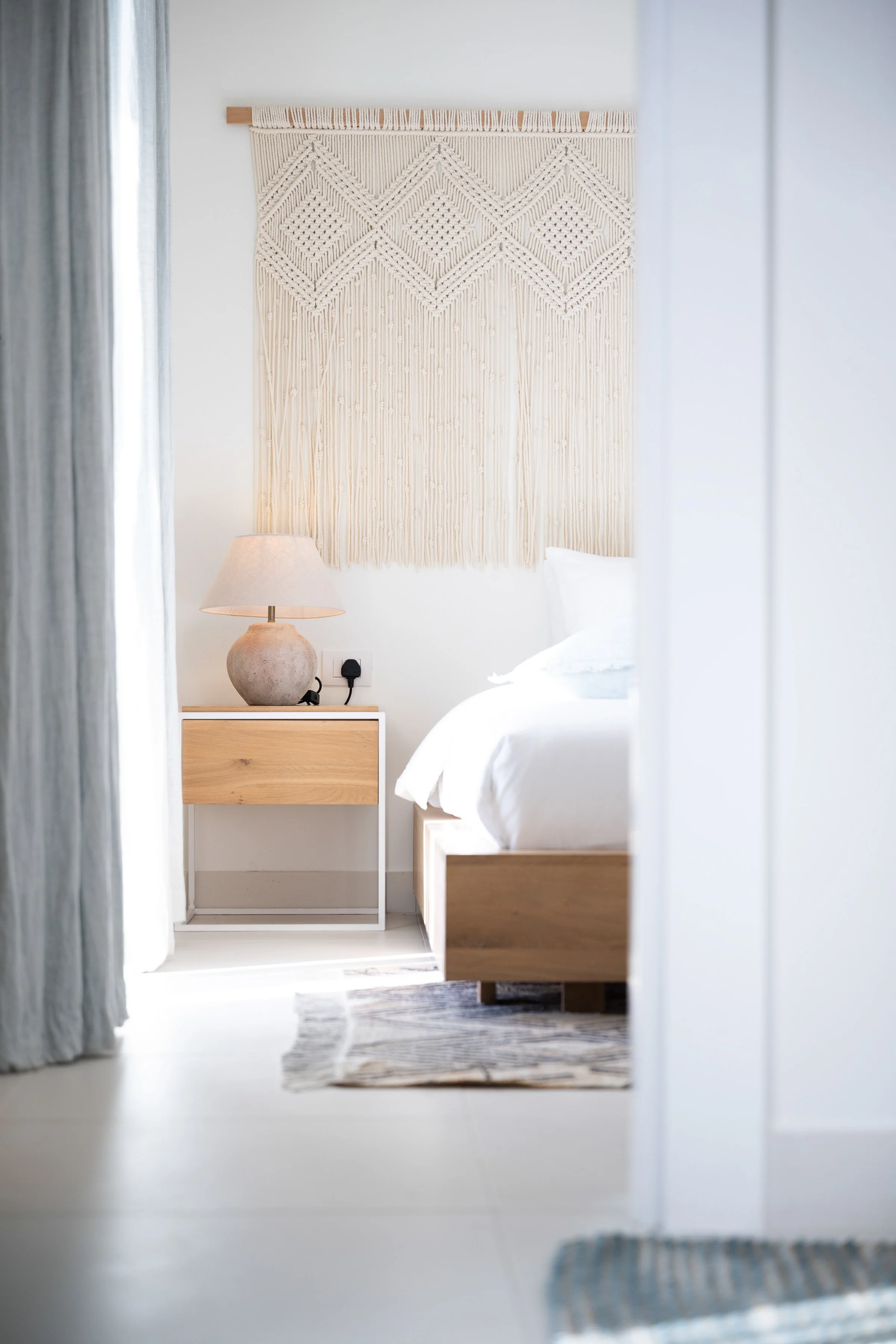 A cozy bedroom with a neutral-toned wall hanging, a bedside table with a lamp, and a neatly made bed with white linens, viewed through an open doorway.