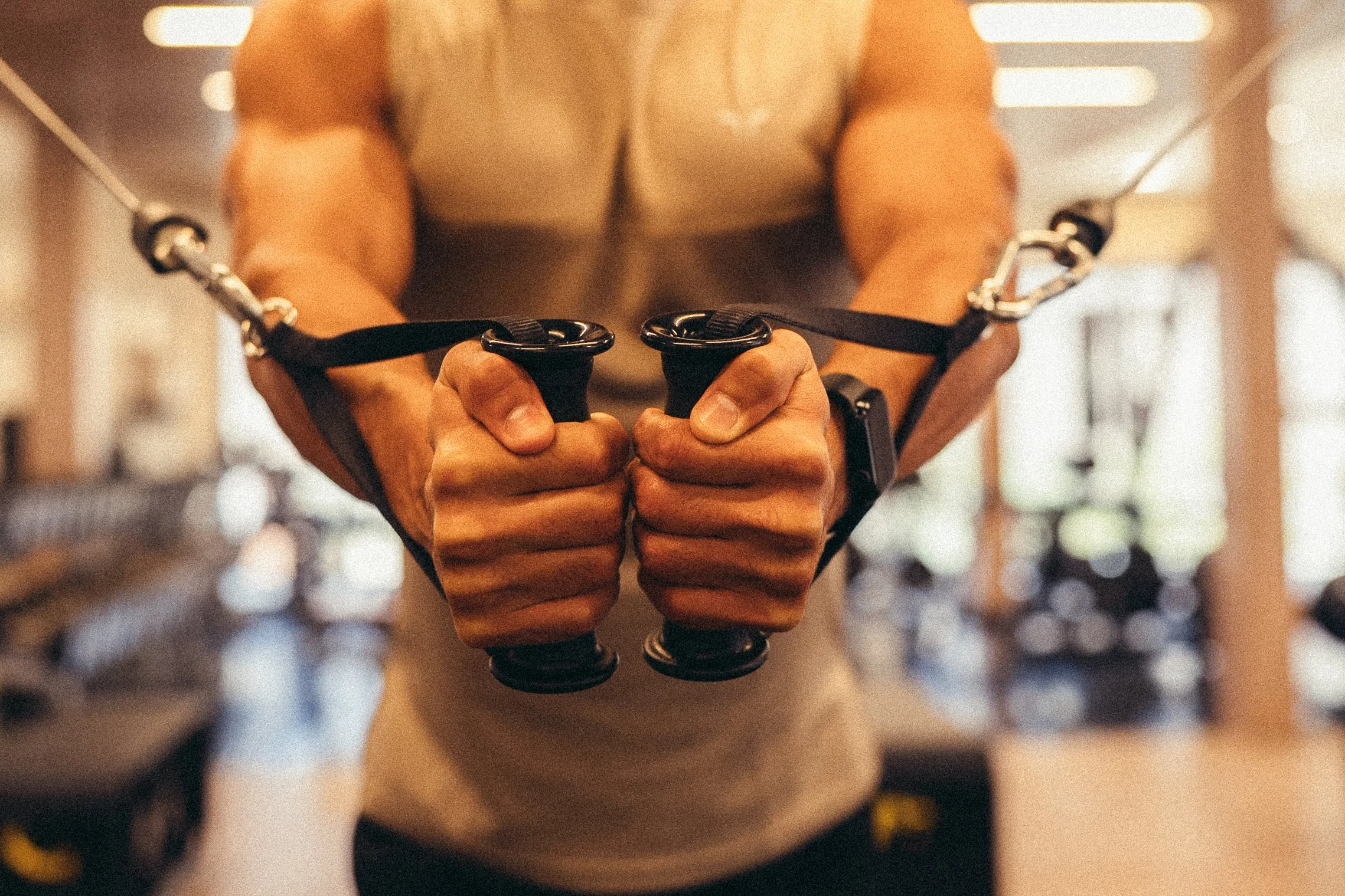 A muscular man in workout attire is gripping handles of a resistance machine at a gym.
