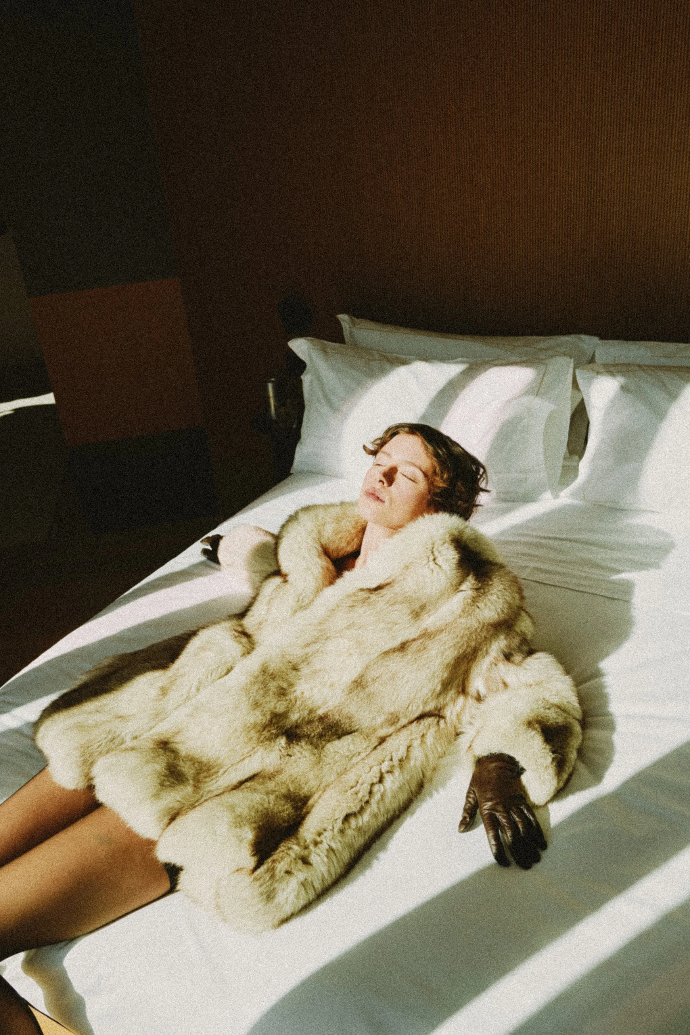 A woman lying on a bed with her eyes closed, wearing a fur coat and gloves, with shadows cast on the bed.