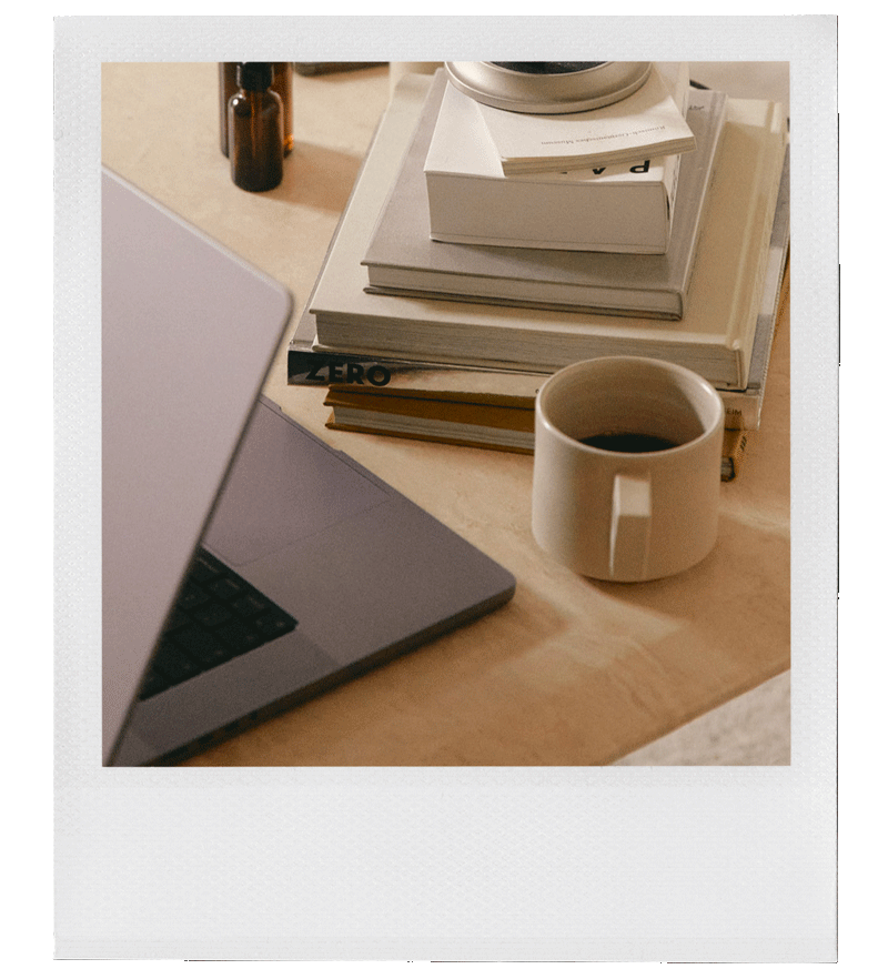 A workspace desk with a laptop, a stack of books and notebooks, a coffee mug, and a small bottle, with part of the laptop and the edge of the desk visible.