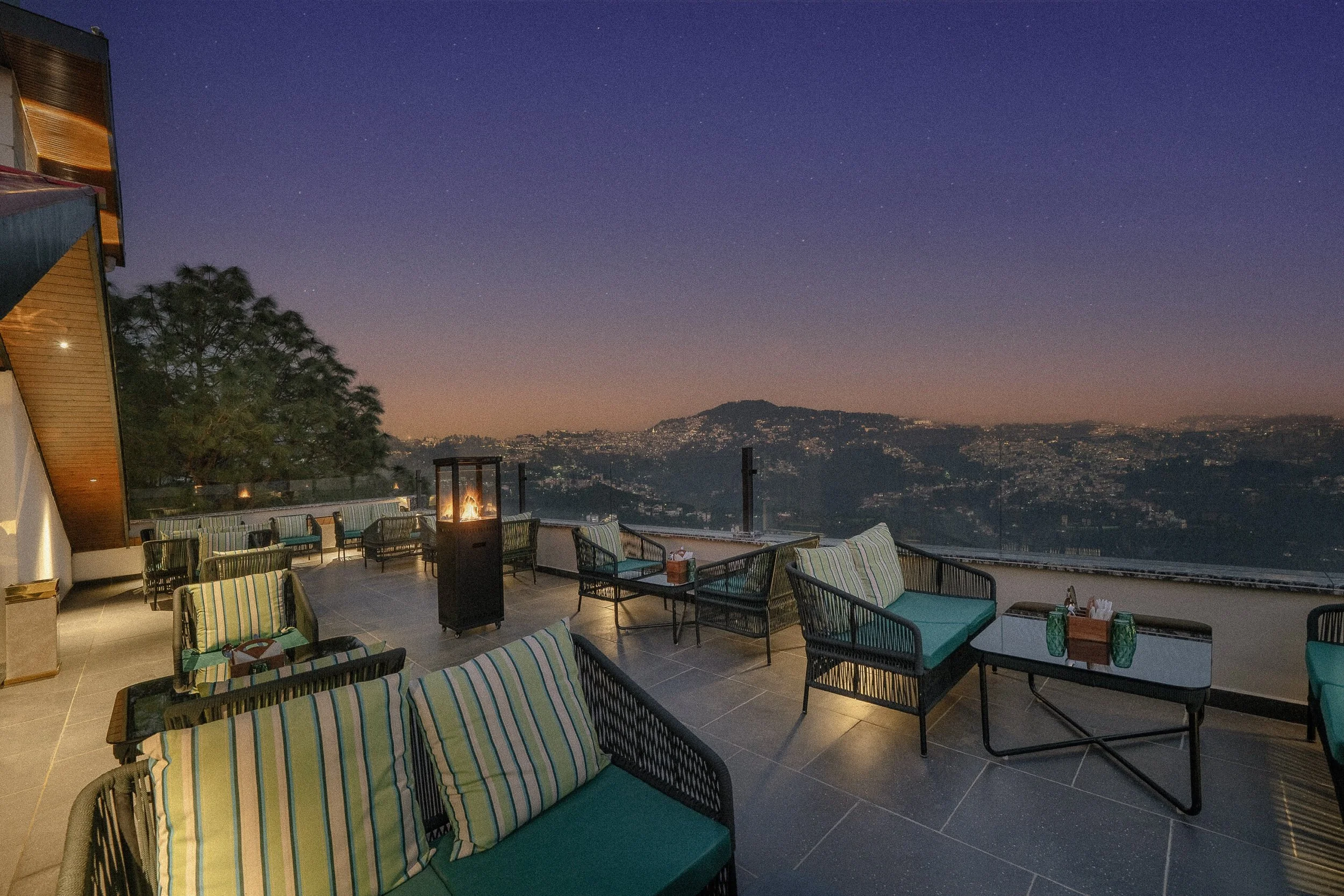 Outdoor rooftop lounge area at dusk with tables, chairs, a fire pit, and a scenic view of hills and city lights in the background.