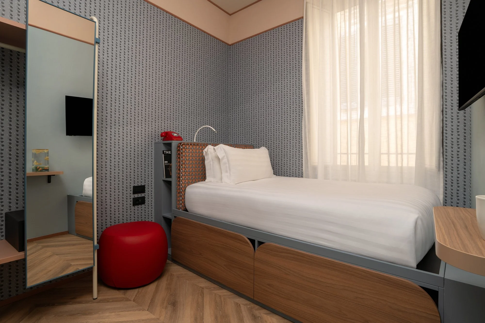 A hotel room with a single bed, a window with sheer curtains, a small bedside table, a Mirror, and a flat-screen TV. There is a red rotary phone and a red stool near the bed, and a fish tank is on a shelf in the corner.