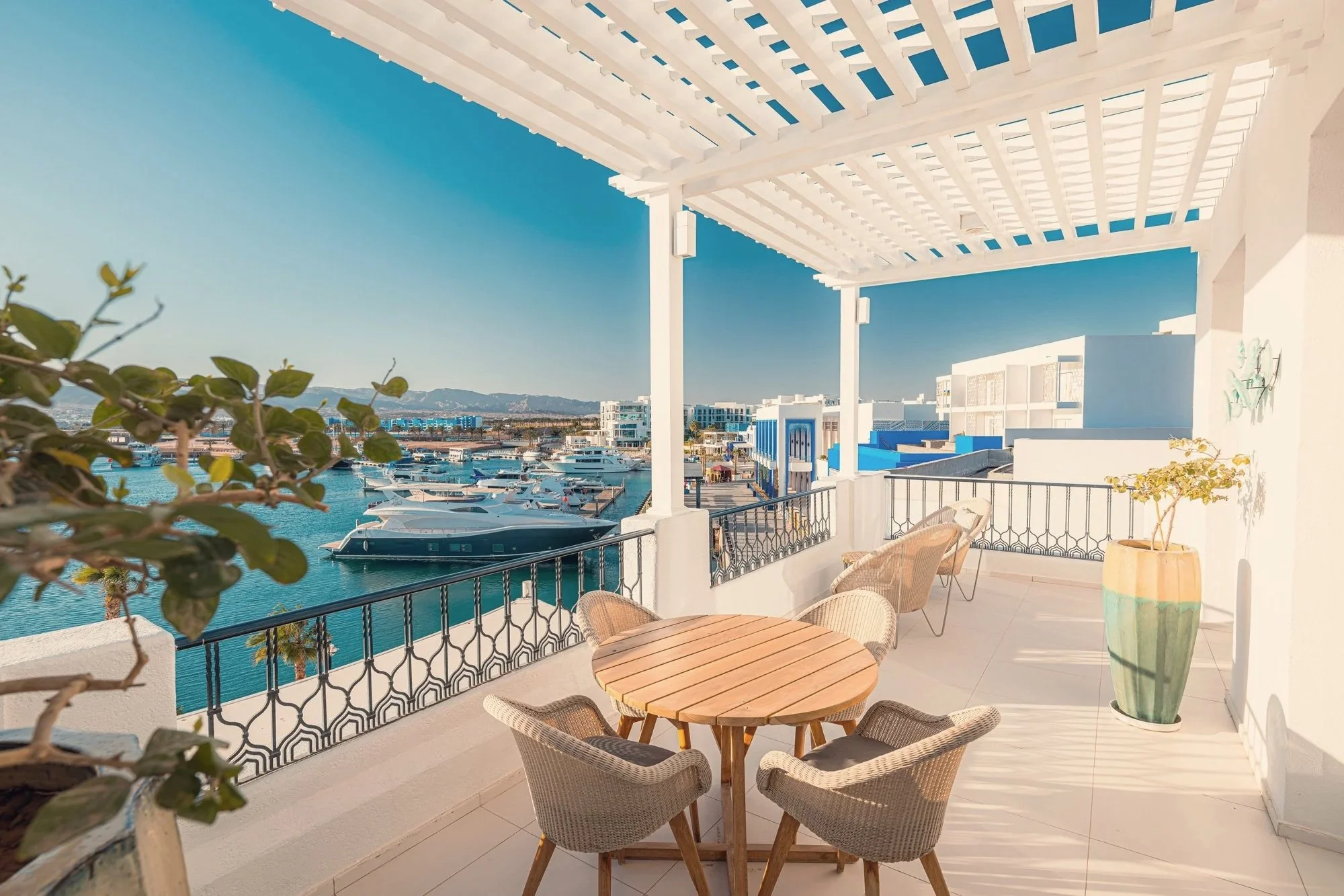 Balcony with a round wooden table and four wicker chairs overlooking a marina with yachts, white buildings with blue accents, and distant mountains under a clear blue sky.