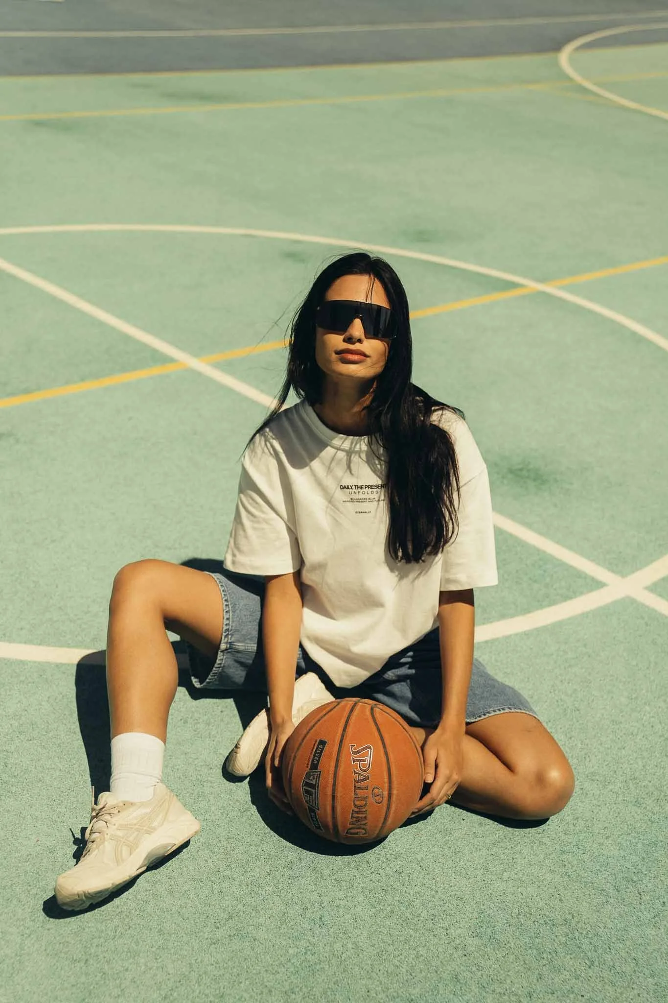 A woman with long black hair, wearing sunglasses, a white T-shirt, denim shorts, and white sneakers, sitting on a basketball court holding a basketball with one leg bent and the other extended.