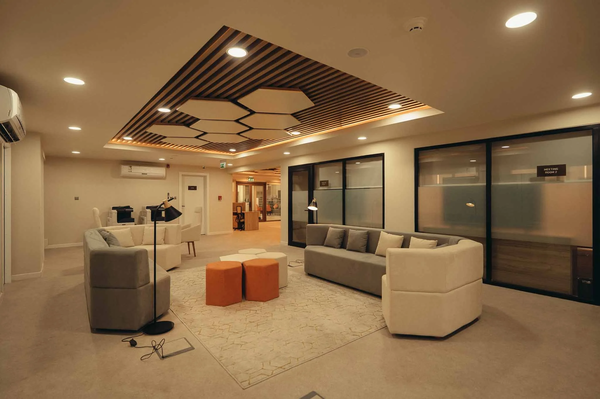 Modern lounge area with beige sofas, colorful ottomans, a patterned rug, and artificial lighting on the ceiling in a hotel or office setting.