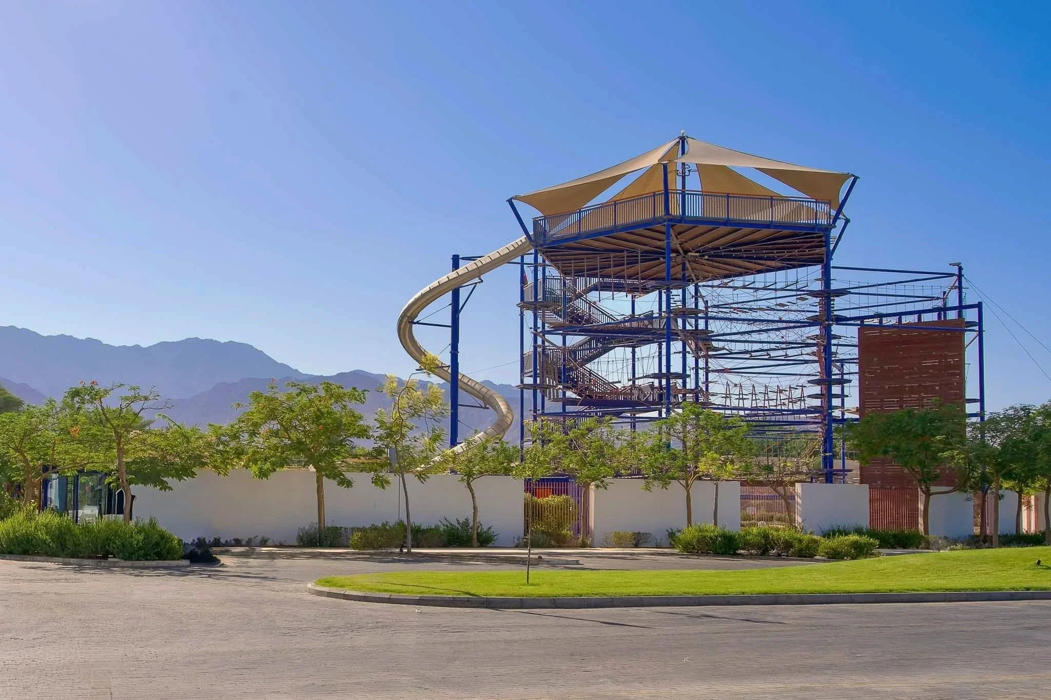 An outdoor water park with a large multi-level play structure, slides, and climbing equipment. Surrounded by trees and mountains in the background, under a clear blue sky.
