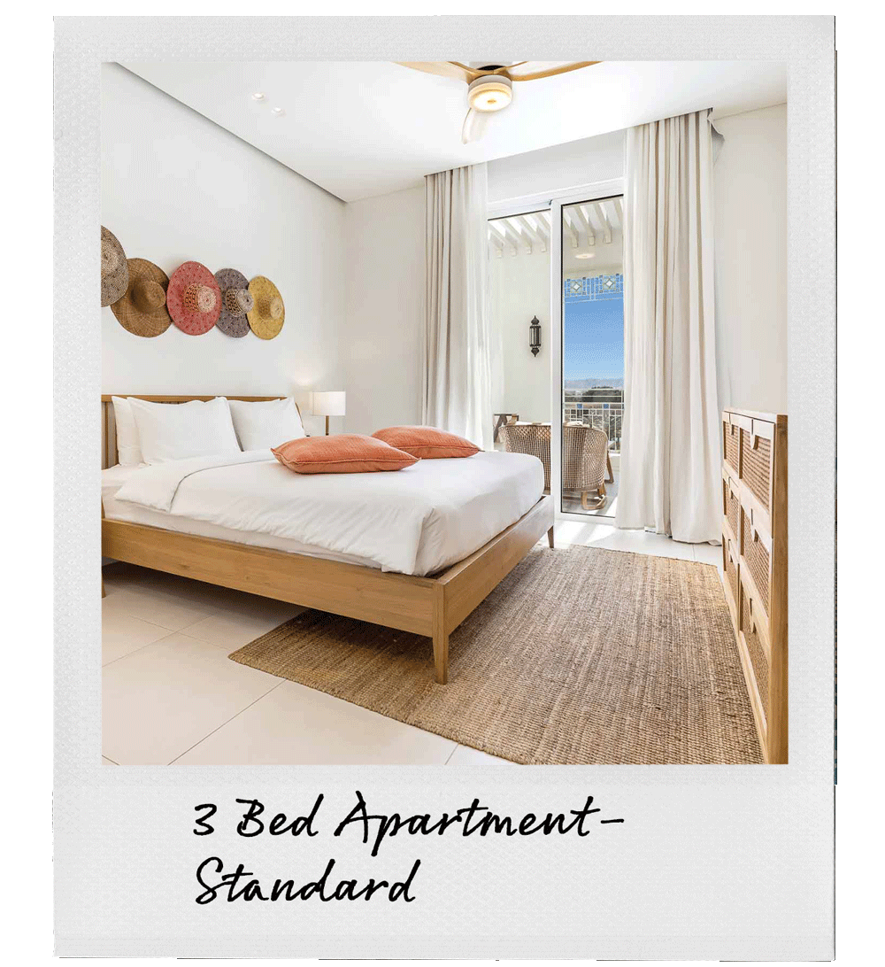 A bedroom with a wooden bed frame, white bedding, orange pillows, a bedside lamp, a woven dresser, and a balcony with outdoor seating and a view of the sky and distant buildings.