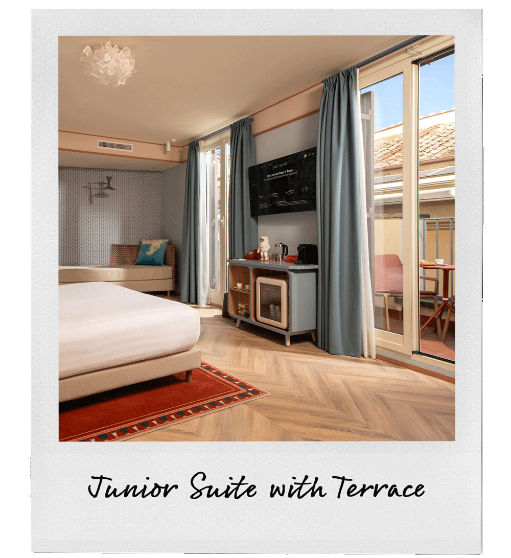 A bedroom in a hotel junior suite with a balcony. The room has a double bed, a small seating area with a chair and a pillow, a TV mounted on the wall, a cabinet with a tea kettle and coffee supplies, and blue curtains. The balcony has a small table and chairs, with a view of neighboring rooftops.