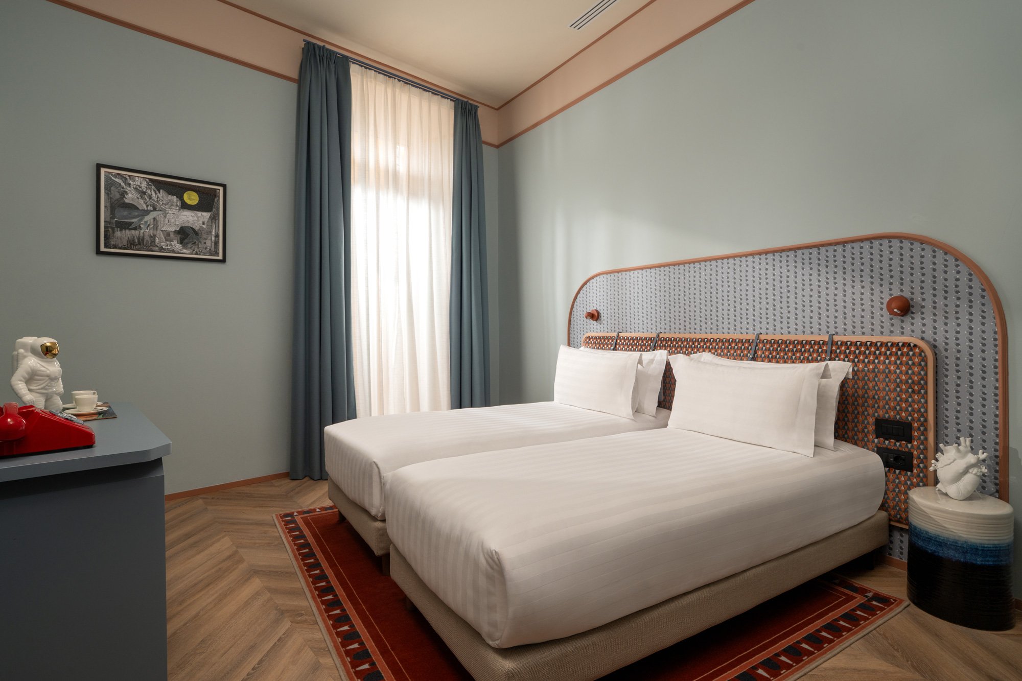 A hotel room with two unmade beds, a decorative headboard, a small bedside table with a white decorative sculpture, a window with blue and white curtains, a framed black and white artwork, and a red rug under the beds.