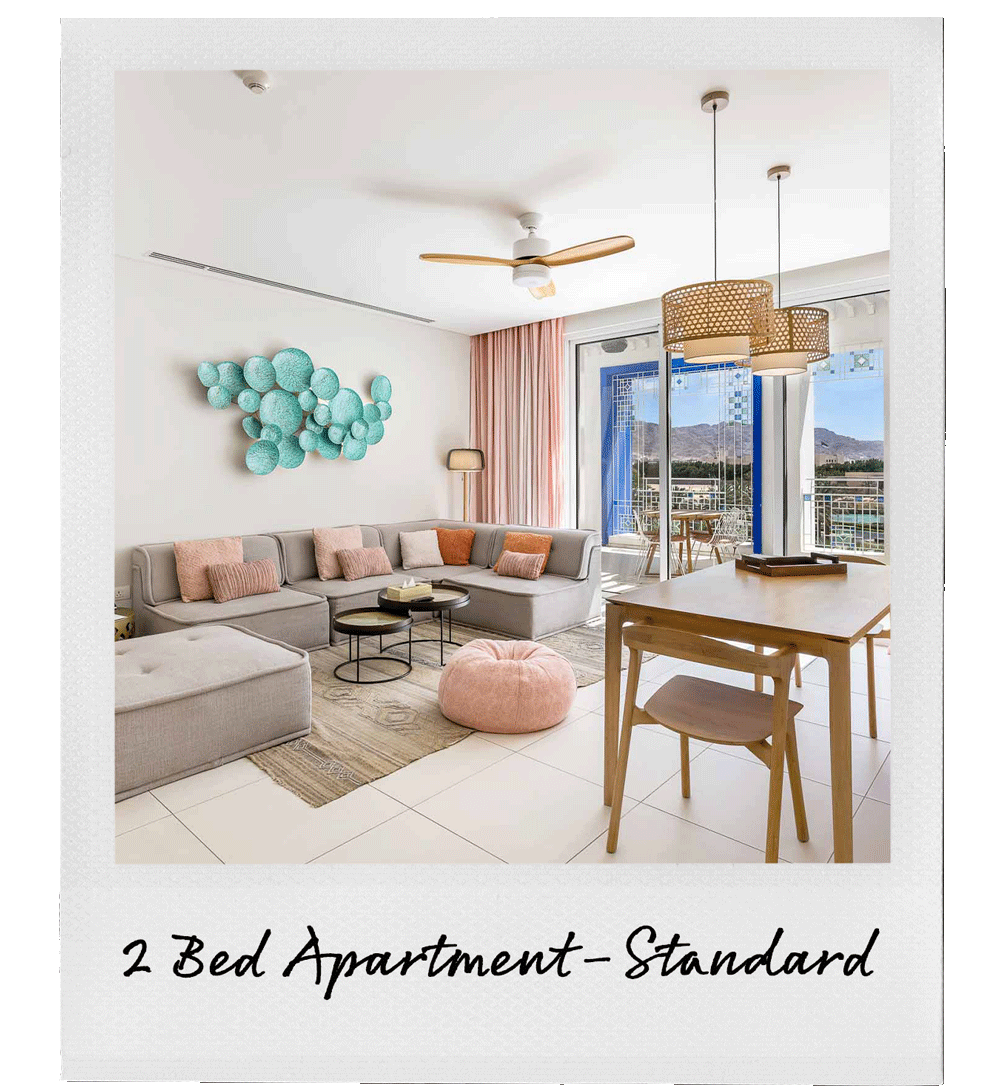 Living room with light-colored sofa, pink and beige throw pillows, a round pink pouf, a wall art sculpture of turquoise spheres, a wooden dining table with a chair, pendant lights, large sliding glass doors leading to a balcony, and mountain views outside.