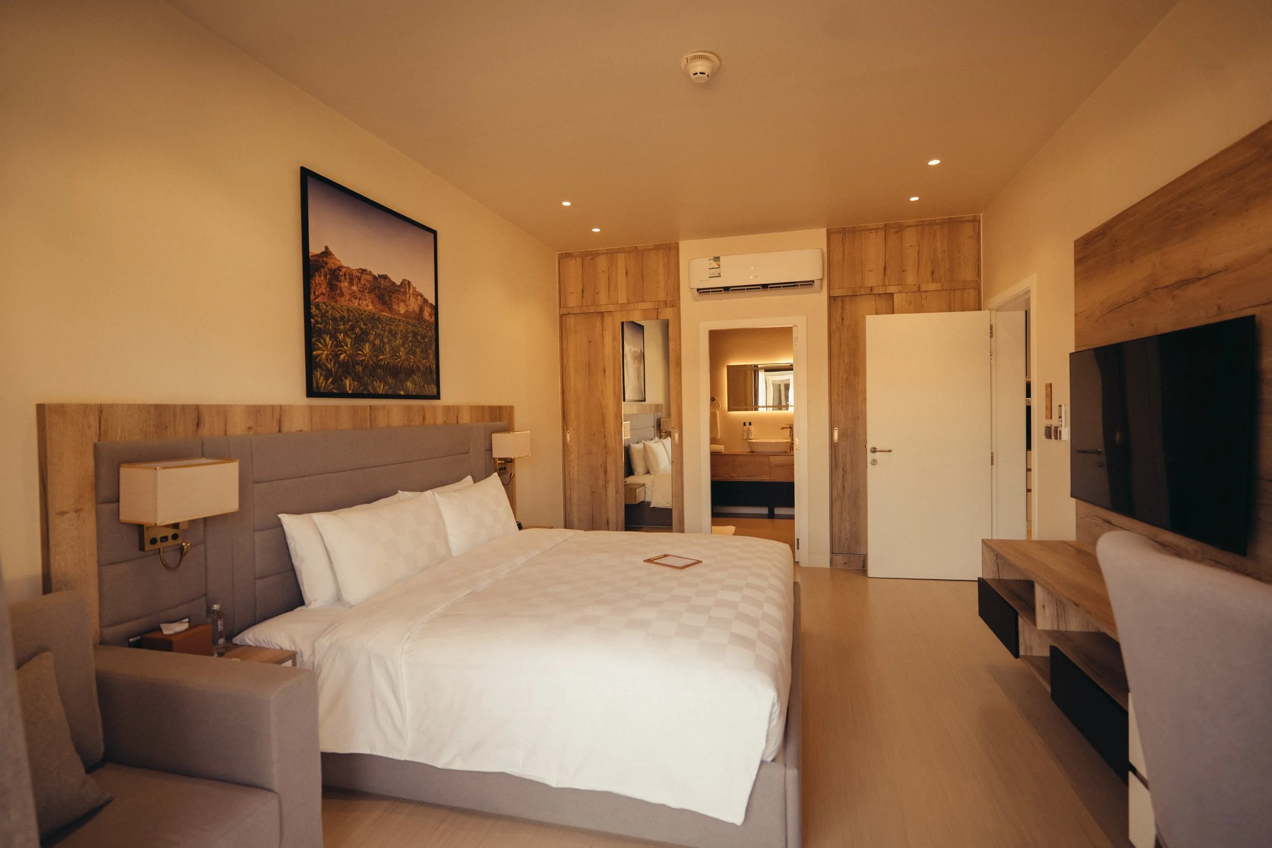 A hotel room with a large bed, wooden accents, a dresser, a wall-mounted TV, and artwork of a mountain landscape. There's a doorway leading to a bathroom with a sink, mirror, and toiletries, and an air conditioning unit above.