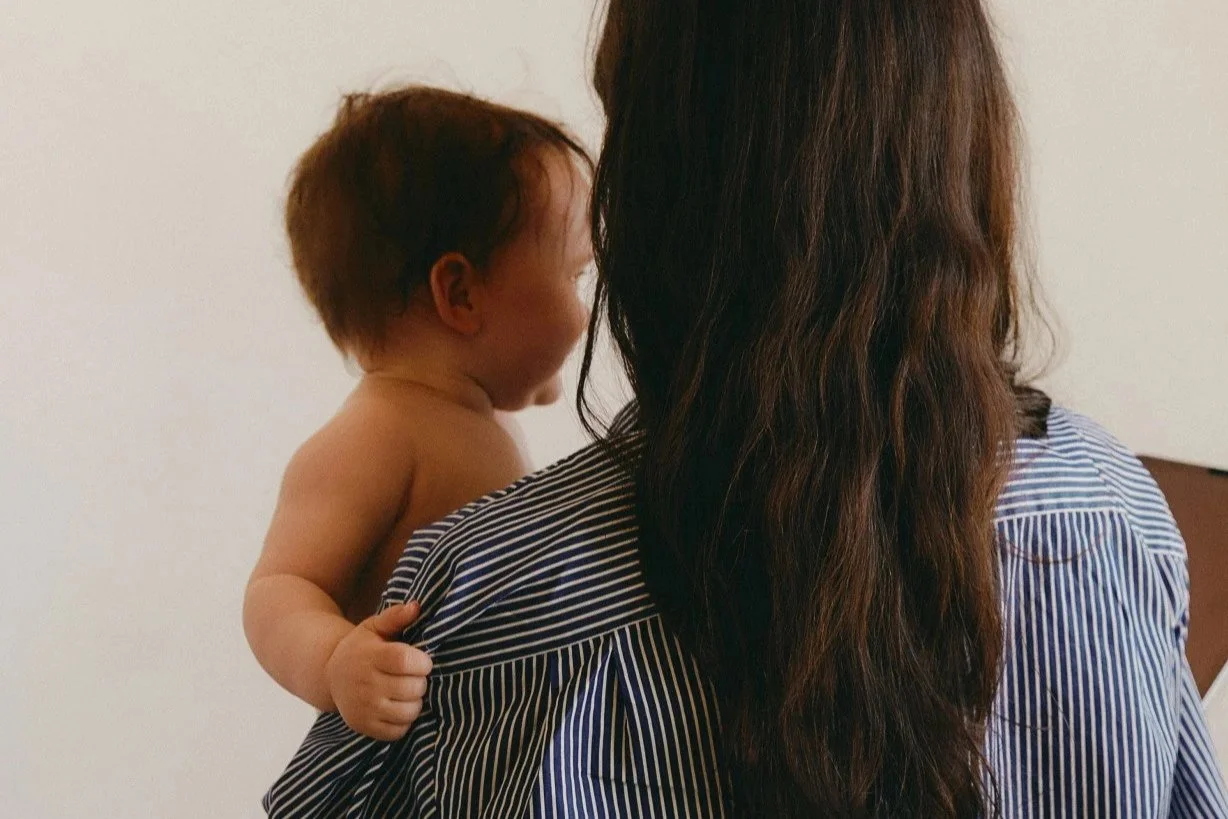 A child hugging a woman, viewed from behind. The child's face is turned toward the woman, and only part of her face is visible. The background is plain white.