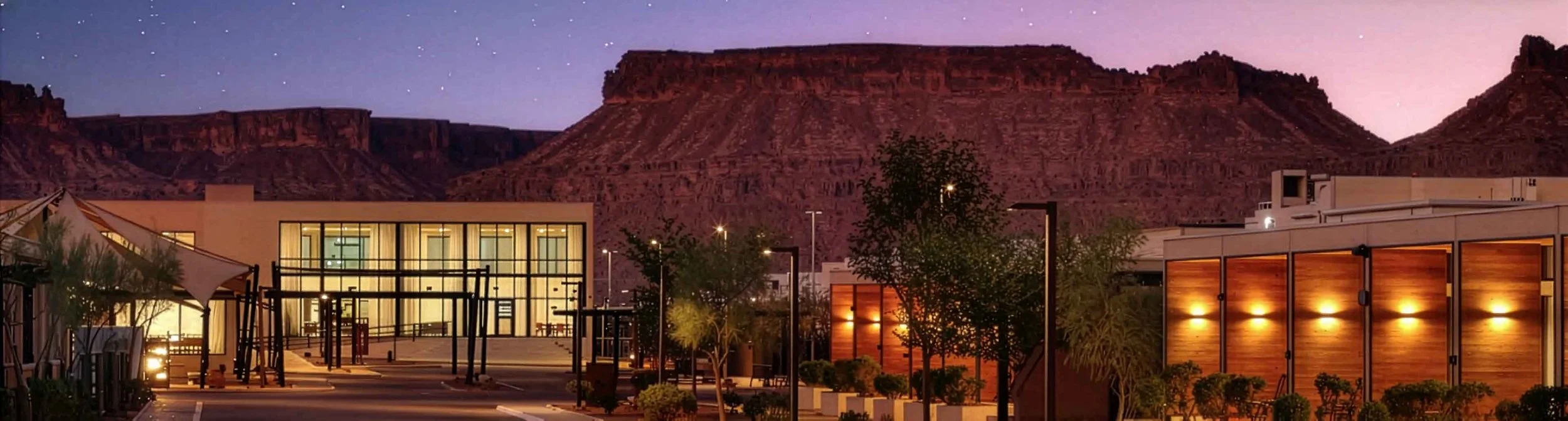Modern building complex with glass windows and wooden panels, trees, and outdoor lighting, with a red rock mesa and starry sky in the background.