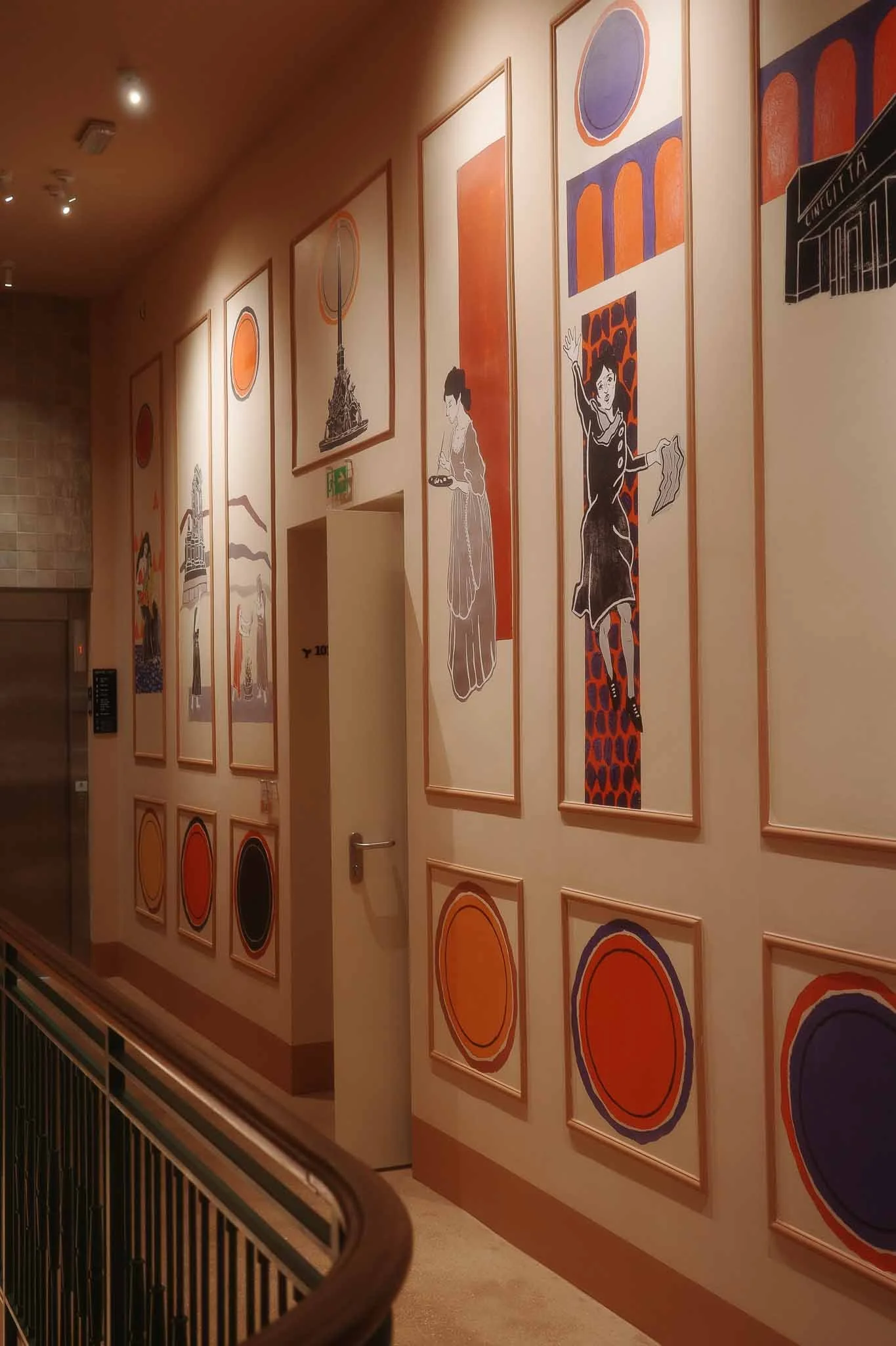 A hallway with a series of framed abstract art pieces on a beige wall. The artwork features circles, geometric shapes, and illustrations of women, with a color palette of orange, purple, and black. A black railing runs along the lower part of the wall, and the ceiling has small spotlights illuminating the art.