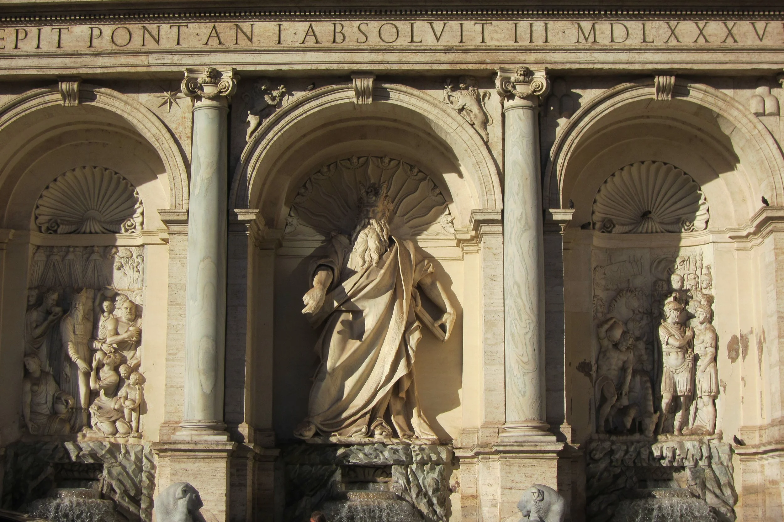 Close-up of the Trevi Fountain in Rome, Italy, showing ornate stone carvings, a central seated figure, and sculptures of figures and animals, with Greek-style columns and Latin inscriptions above.