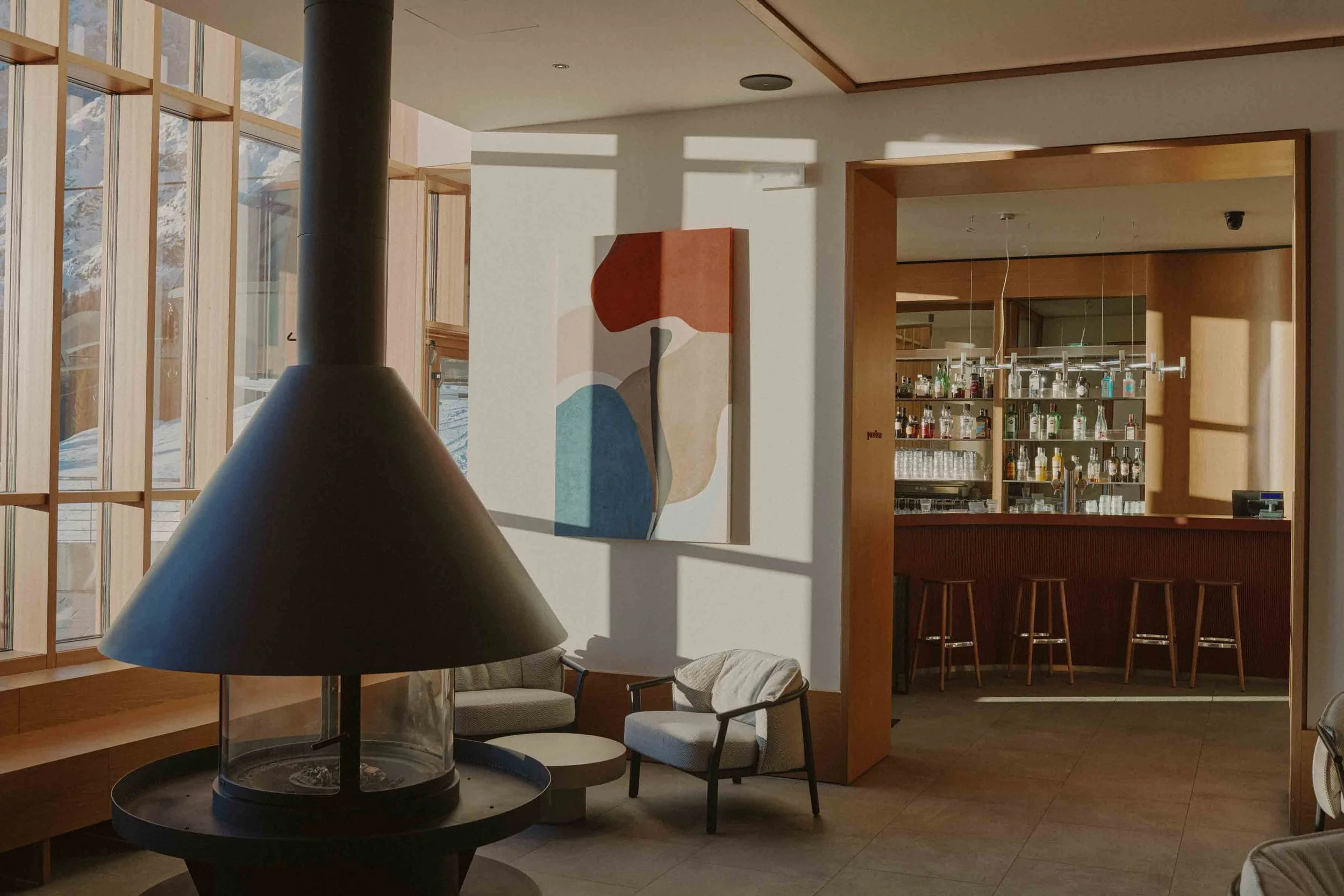 Interior view of a cozy space with wooden accents, a bar area with bottles on shelves, seating with chairs and a sofa, and sunlight streaming through large windows casting shadows.