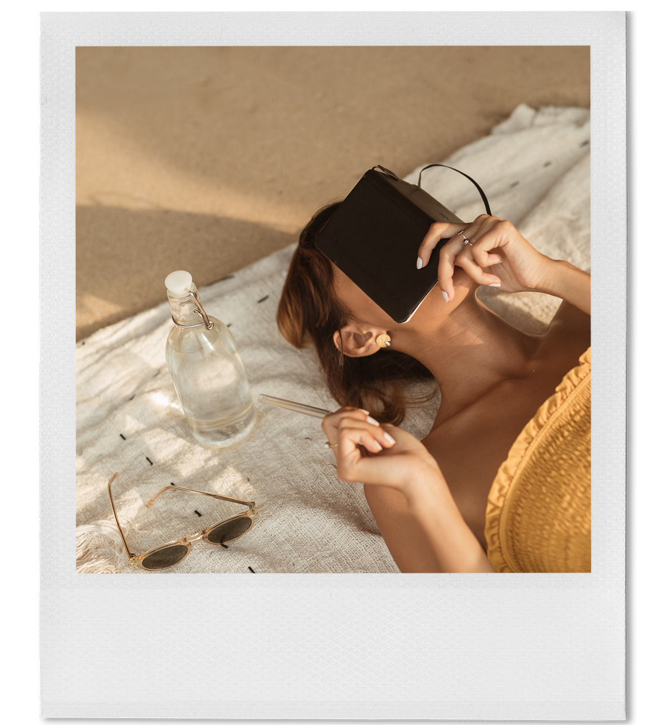 A woman with brown hair and earrings lying on a bed or blanket, holding a closed tablet over her face, with sunglasses and a water bottle next to her.