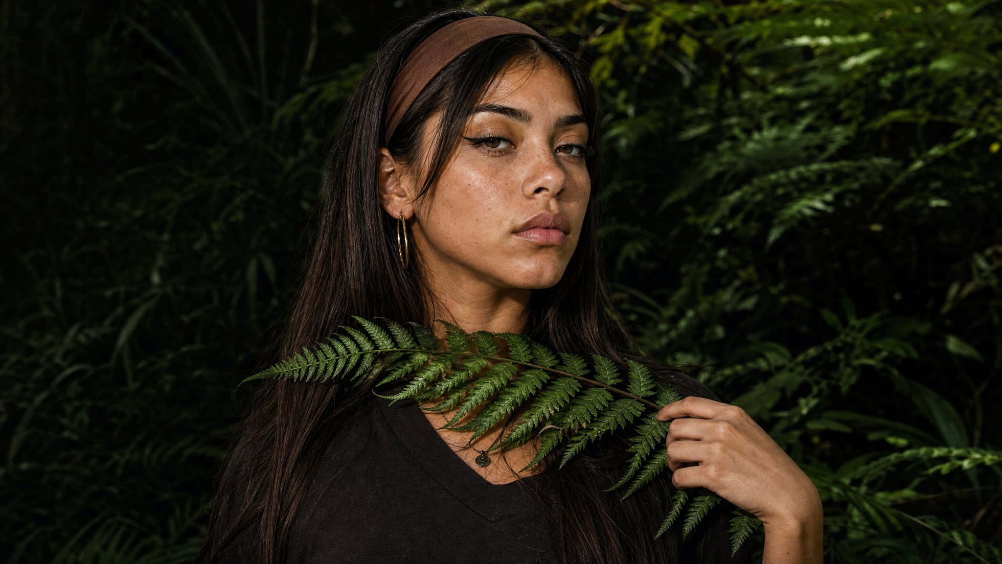 Junge Frau mit dunklen Haaren, braunem Kopfband, großen goldenen Ohrringen und dunklem Shirt steht in einem grünen Dschungel und hält ein Farnblatt vor den Hals.