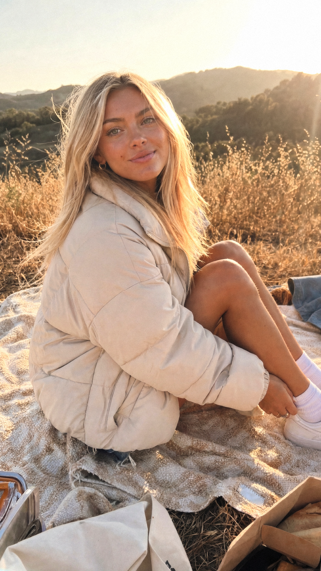 Junge Frau sitzt auf einer Decke im Freien bei Sonnenuntergang, umgeben von Natur und Bergen im Hintergrund, trägt einen cremefarbenen Pufferjacke, lächelt.