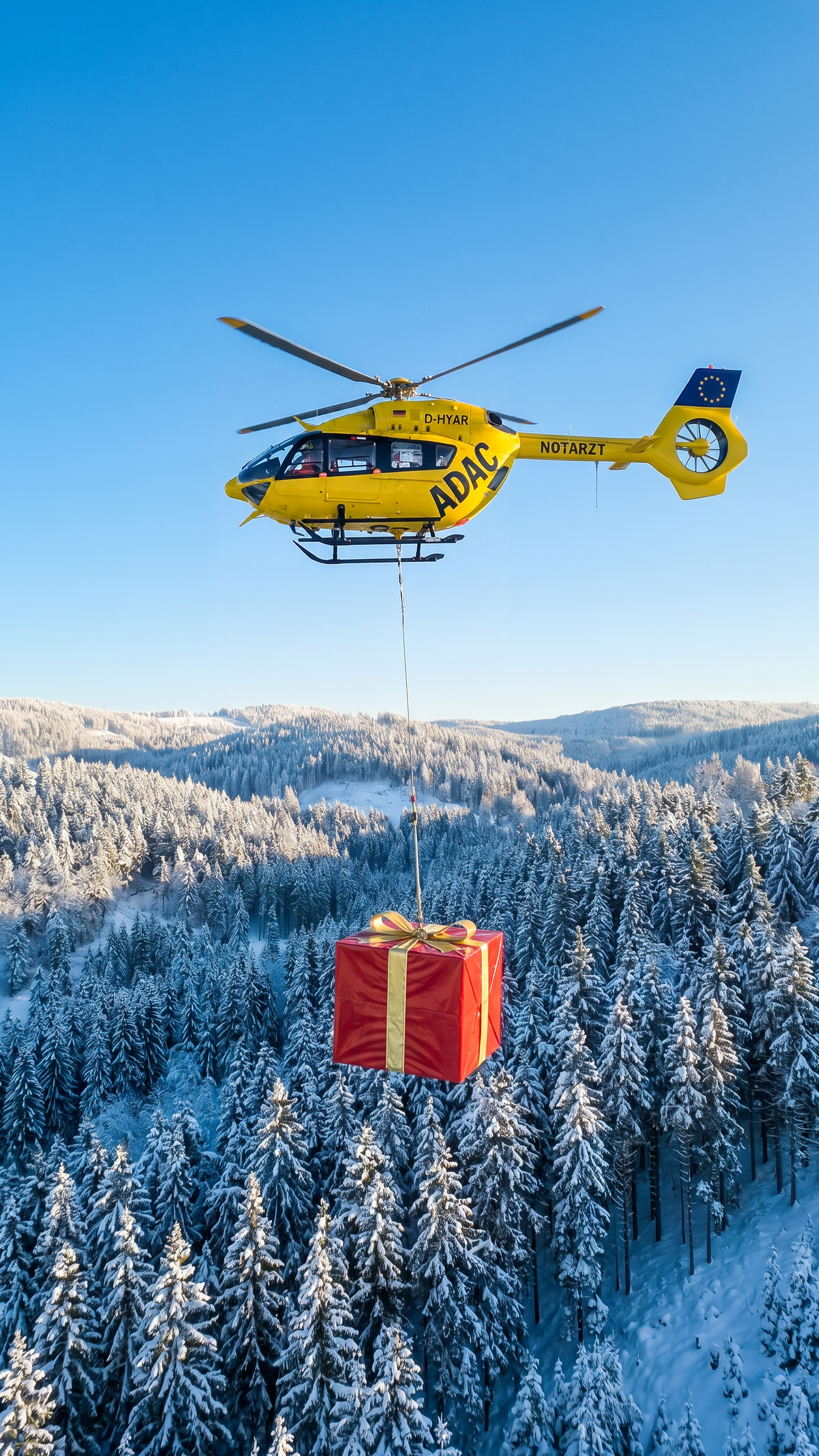 Ein Rettungshubschrauber hantiert mit einer Seilwinde, die ein rotes Geschenk mit gelber Schleife trägt, über einer verschneiten Winterlandschaft.