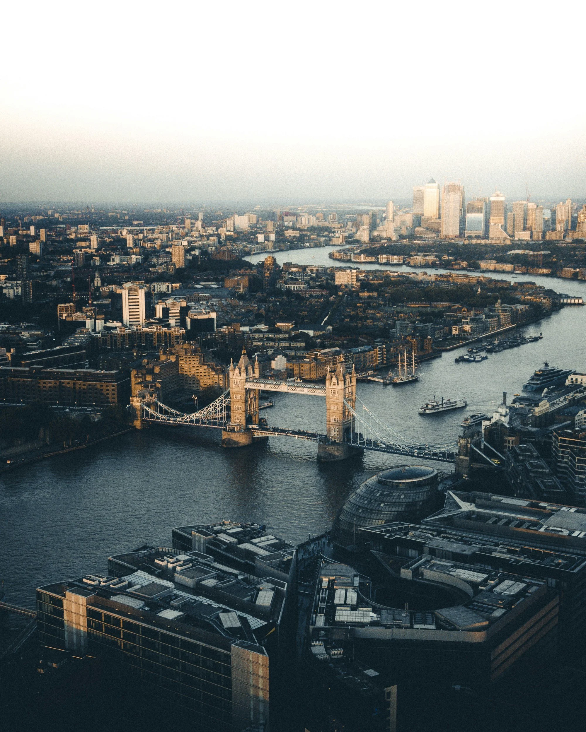Aerial view of a city skyline featuring skyscrapers, a river with boats and bridges, including the Tower Bridge in London, England, during sunset.