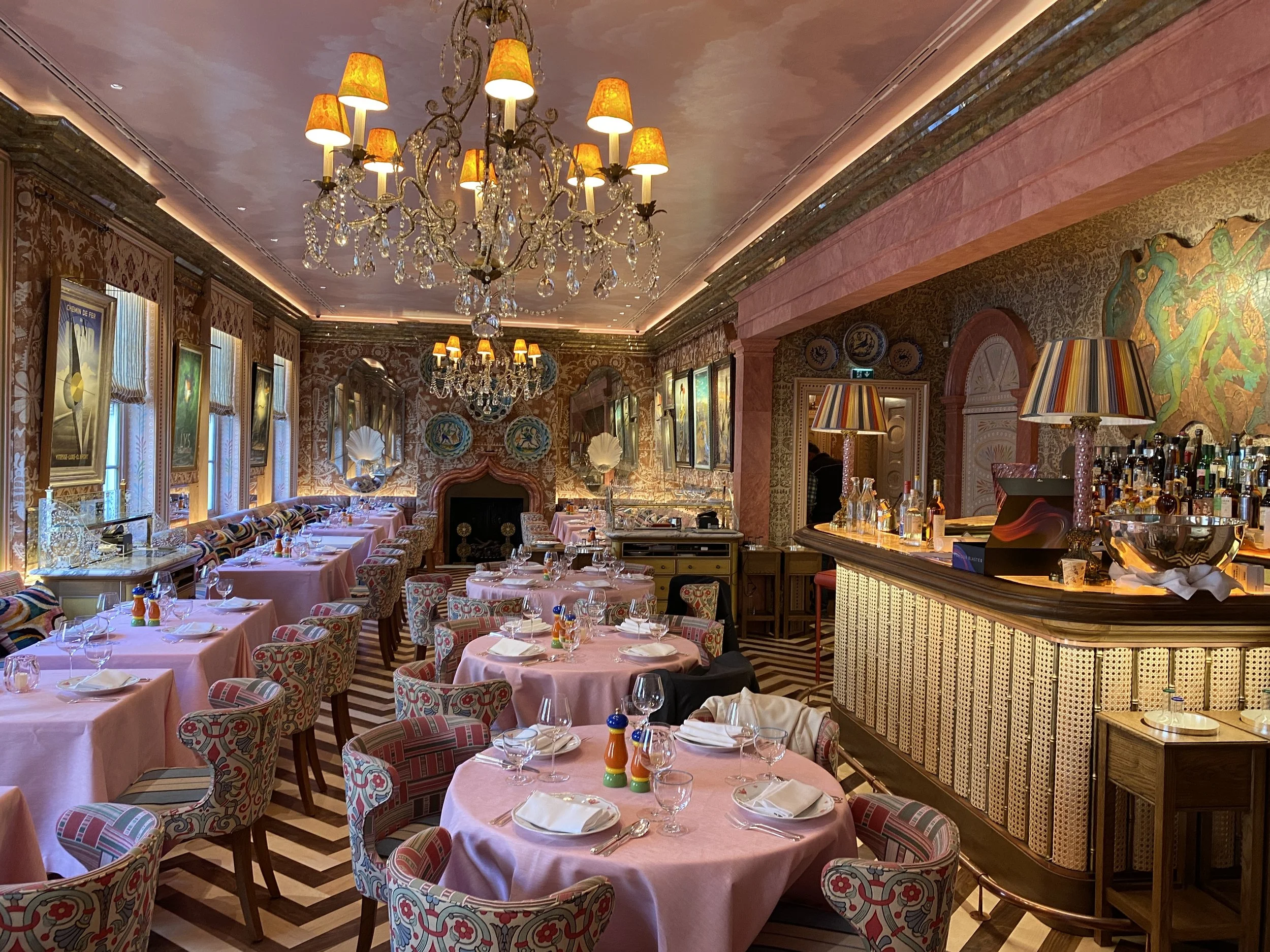 An elegant restaurant interior with pink tablecloths, patterned chairs, chandeliers, colorful lamps, and decorated walls with artwork and wallpaper.