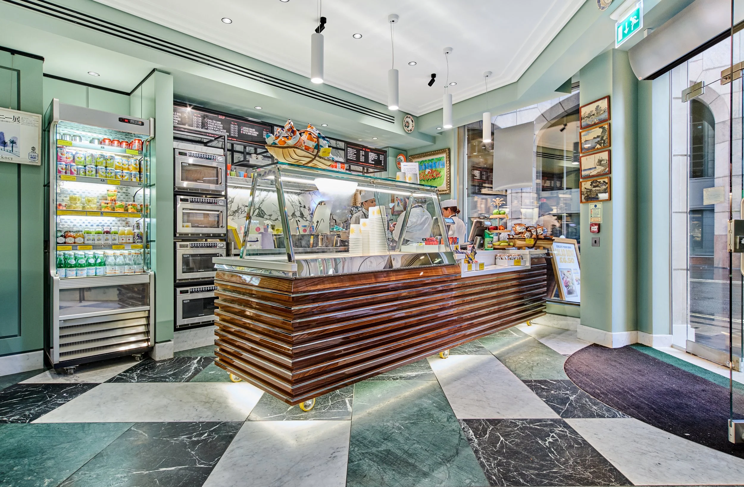 Interior of a cafe or small restaurant with a counter and staff preparing food, shelves with packaged goods, a refrigerator, and a menu board, decorated with paintings and situated by a glass door with an exit sign.