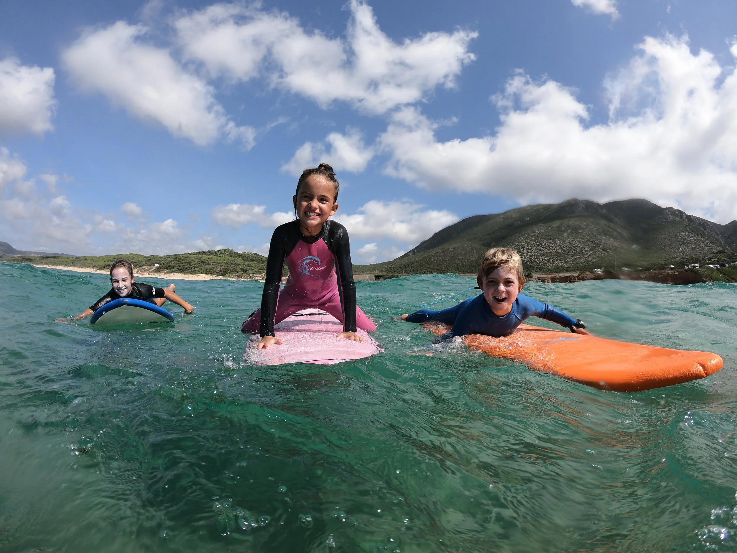 Bambini felici che fanno surf su onde nell'oceano con montagne sullo sfondo e un cielo nuvoloso.
