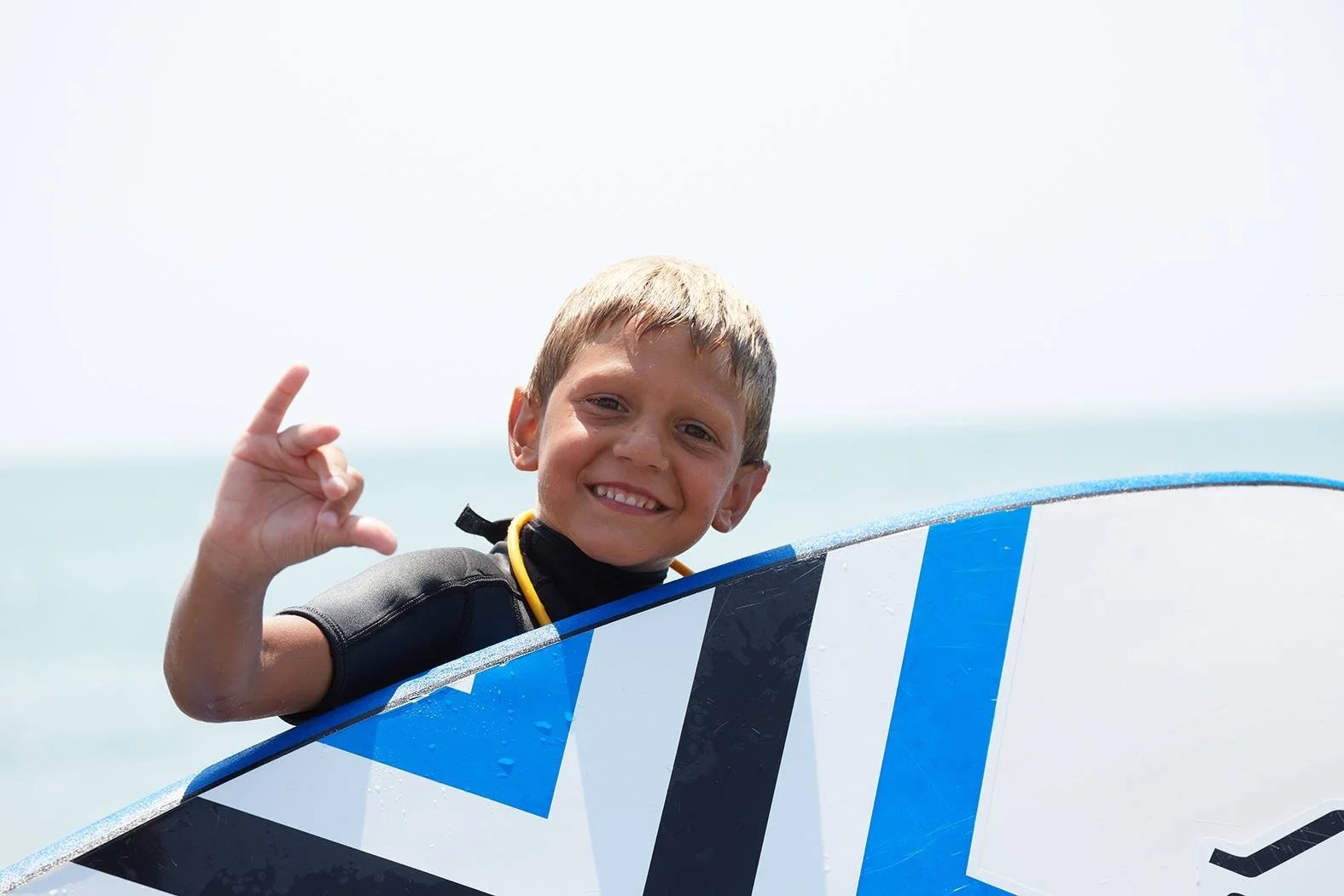 Un bambino sorridente tiene una tavola da surf con un simbolo di pace e fa il gesto dello shaka con la mano.