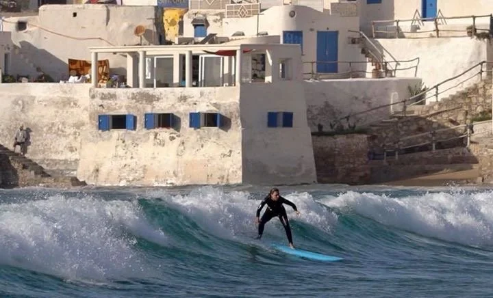 Una surfista che cavalca le onde vicino a edifici bianchi con dettagli blu, in un ambiente costiero mediterraneo.
