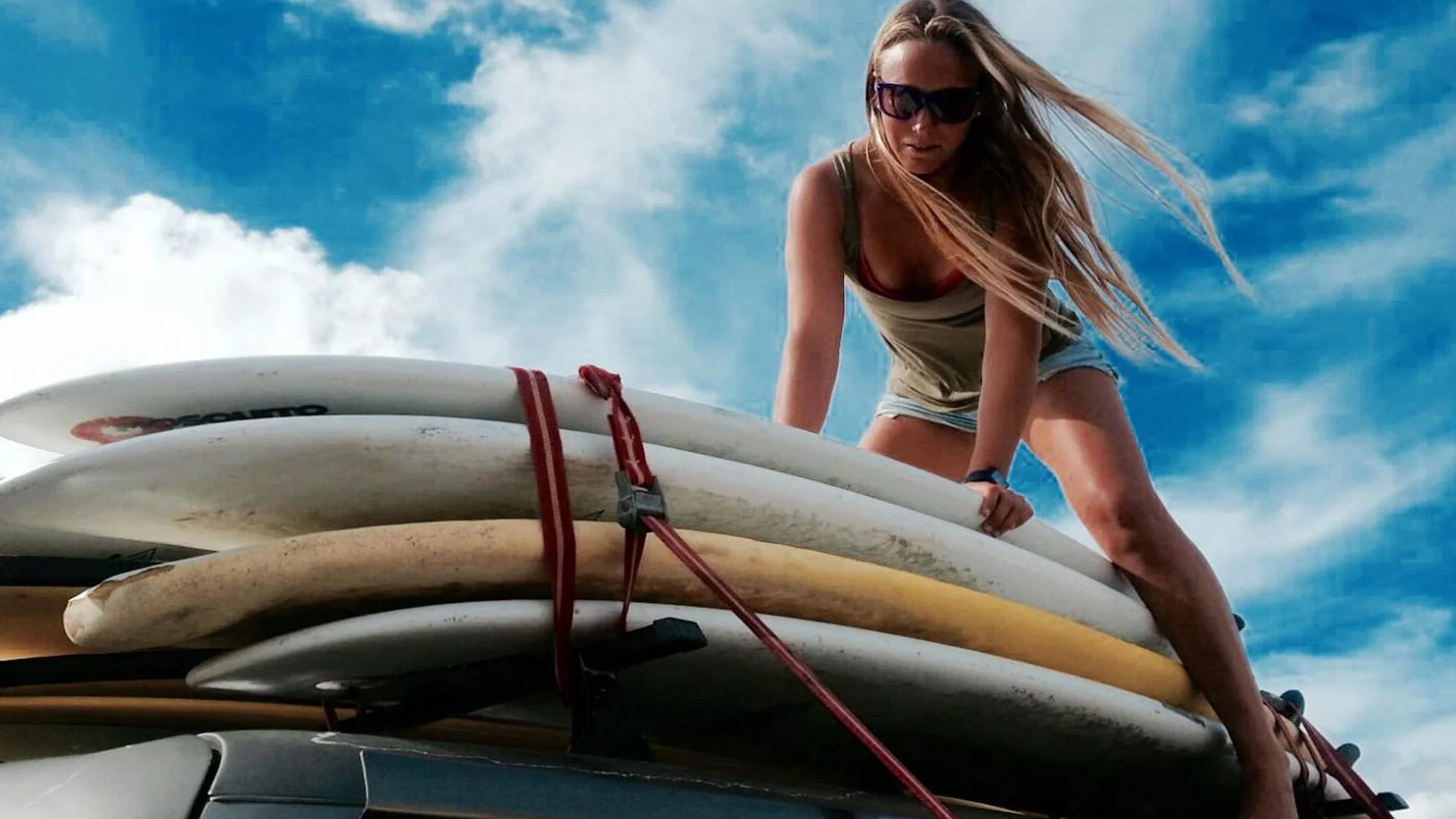 Una donna con occhiali da sole e capelli lunghi si sta arrampicando su delle tavole da surf attaccate su un'auto, il cielo è blu con alcune nuvole.