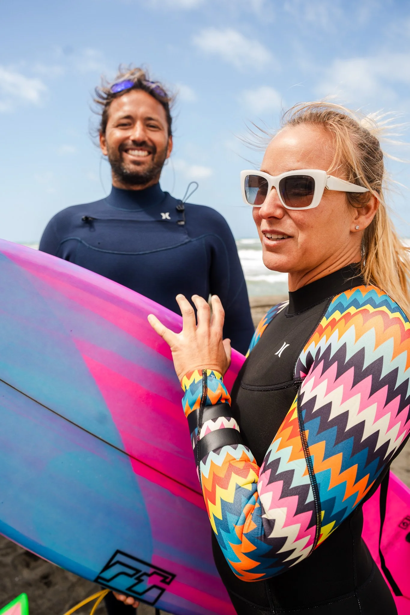 Due persone sulla spiaggia, una donna in primo piano con occhiali da sole e una maglietta colorata, una donna dietro con una tavola da surf rosa e blu, entrambi sorridenti.