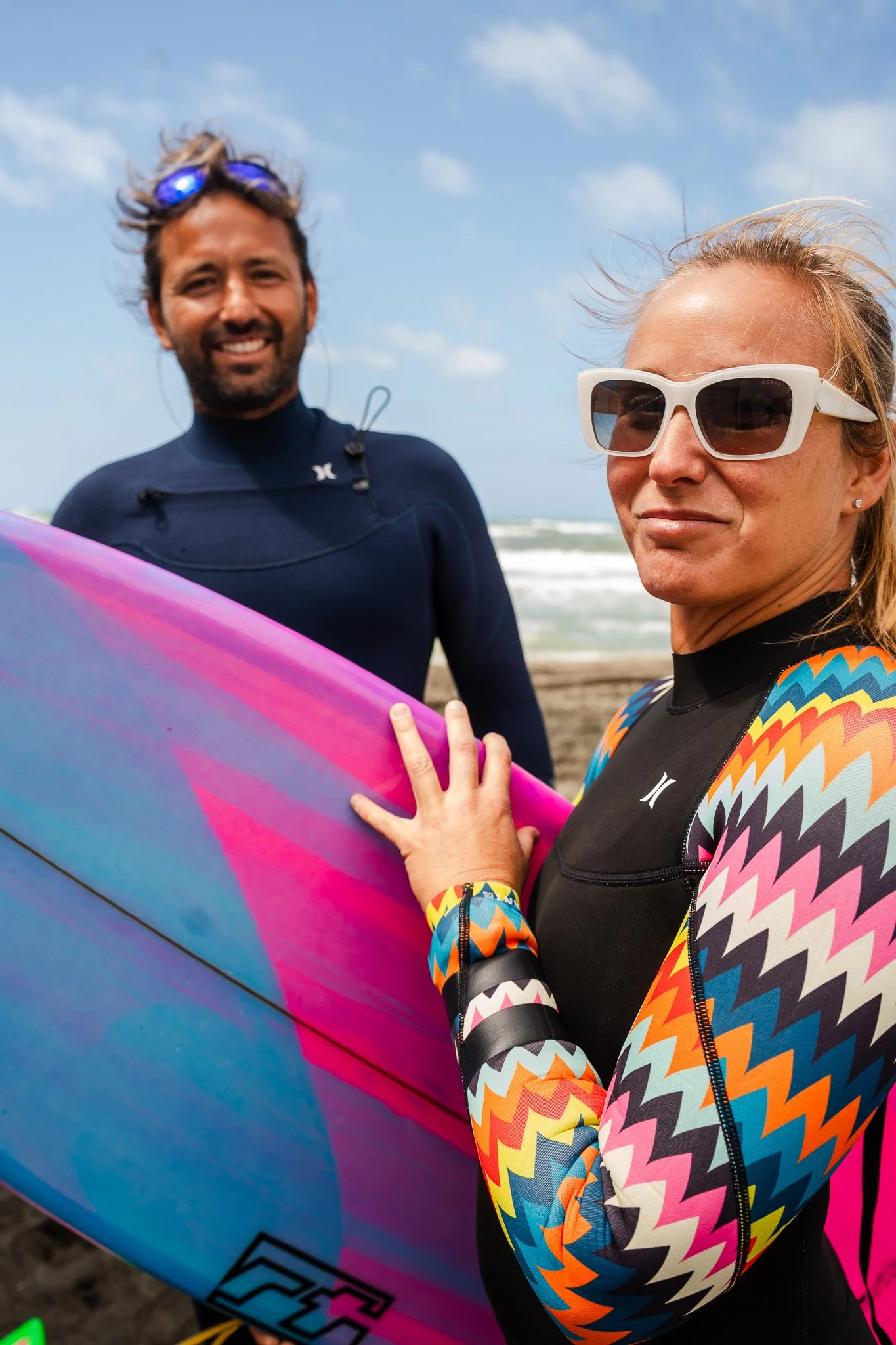 Due persone sulla spiaggia, una donna con occhiali da sole e abbigliamento colorato, un uomo con muta da surf e occhiali di nuoto appoggiati sulla fronte, che tengono una tavola da surf con sfumature rosa, blu e viola.