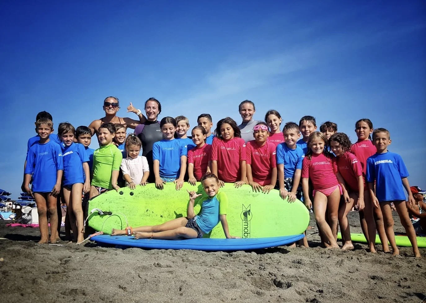 Gruppo di bambini e istruttori sulla spiaggia con tavole da surf colorate, sorridenti e in costume da bagno, sotto un cielo blu.