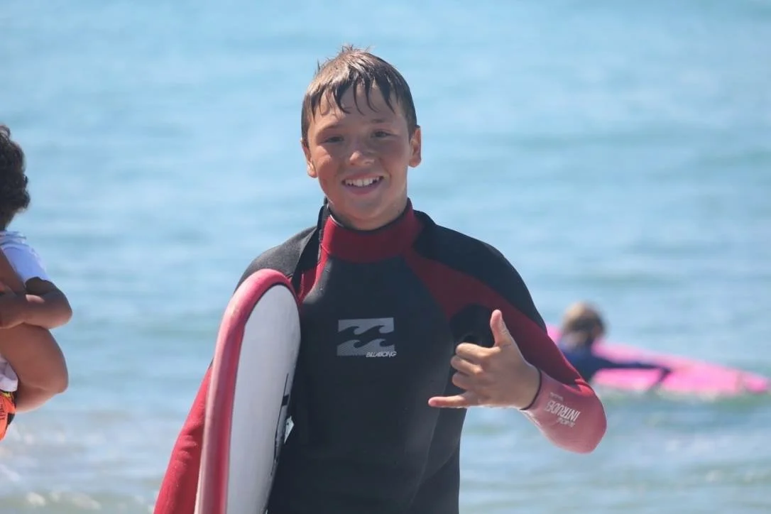 Un ragazzo sorridente con muta da surf e tavola da surf, che fa il gesto della 'rock on' sulla spiaggia con il mare sullo sfondo.