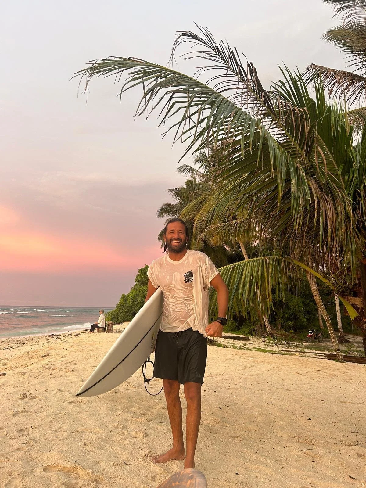 Uomo con maglietta beige e pantaloncini neri che tiene una tavola da surf sulla spiaggia con palme e mare al tramonto