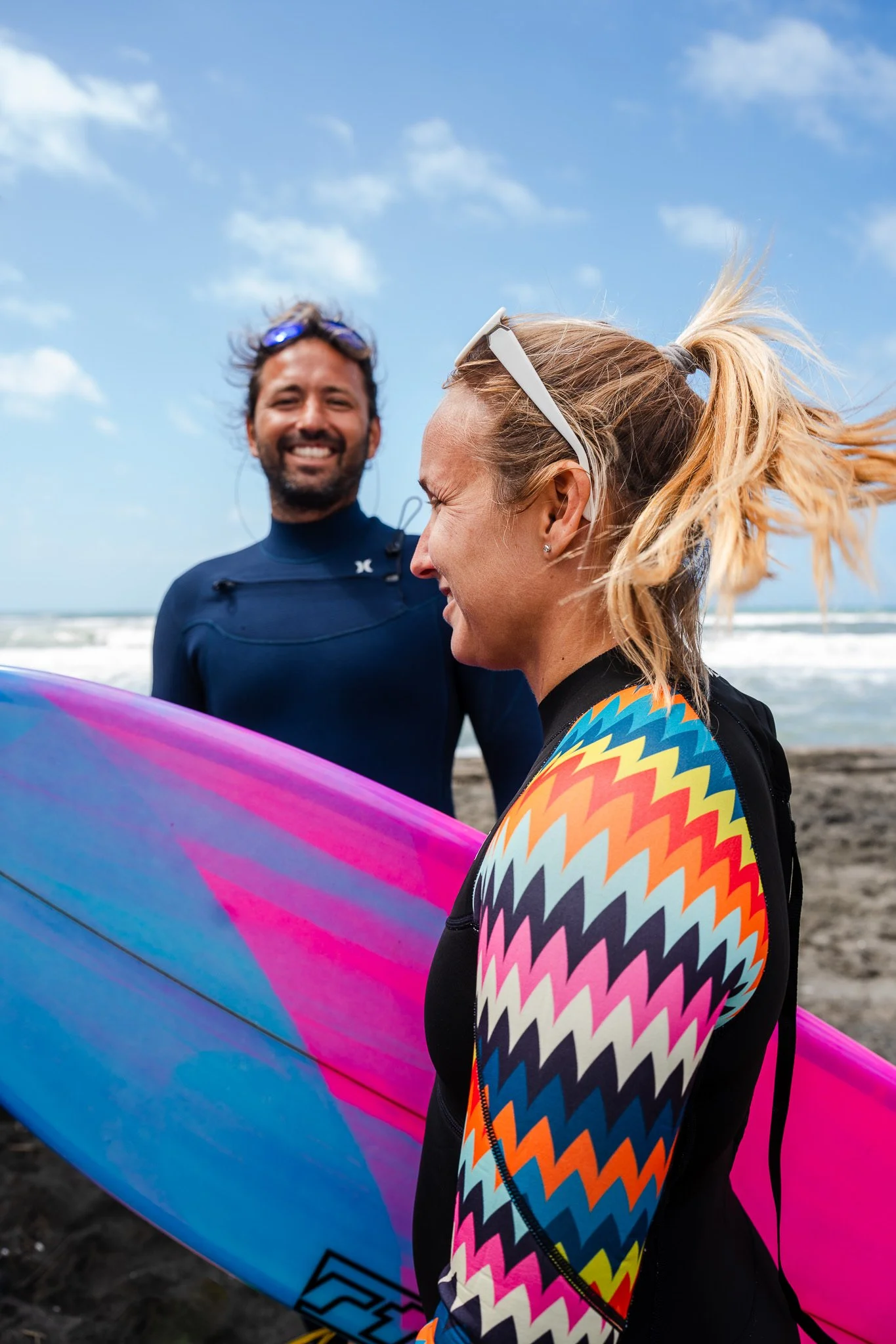 Due persone sorridenti sulla spiaggia, una con una tavola da surf colorata e una con un costume da surf con motivo zigzag, vento tra i capelli e cielo blu con nuvole