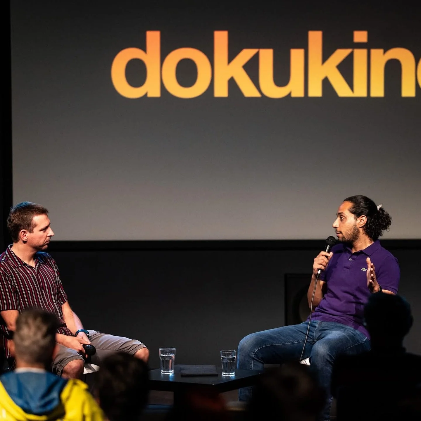 Two men are on stage at a discussion or presentation event. The man on the left has short hair, is wearing a striped shirt, beige shorts, and is seated holding a microphone. The man on the right has long, curly hair tied back, a beard, and is wearing a purple polo shirt and jeans, speaking into a microphone. The background features a large screen with the partially visible word "doku" in yellow letters against a gray gradient background.