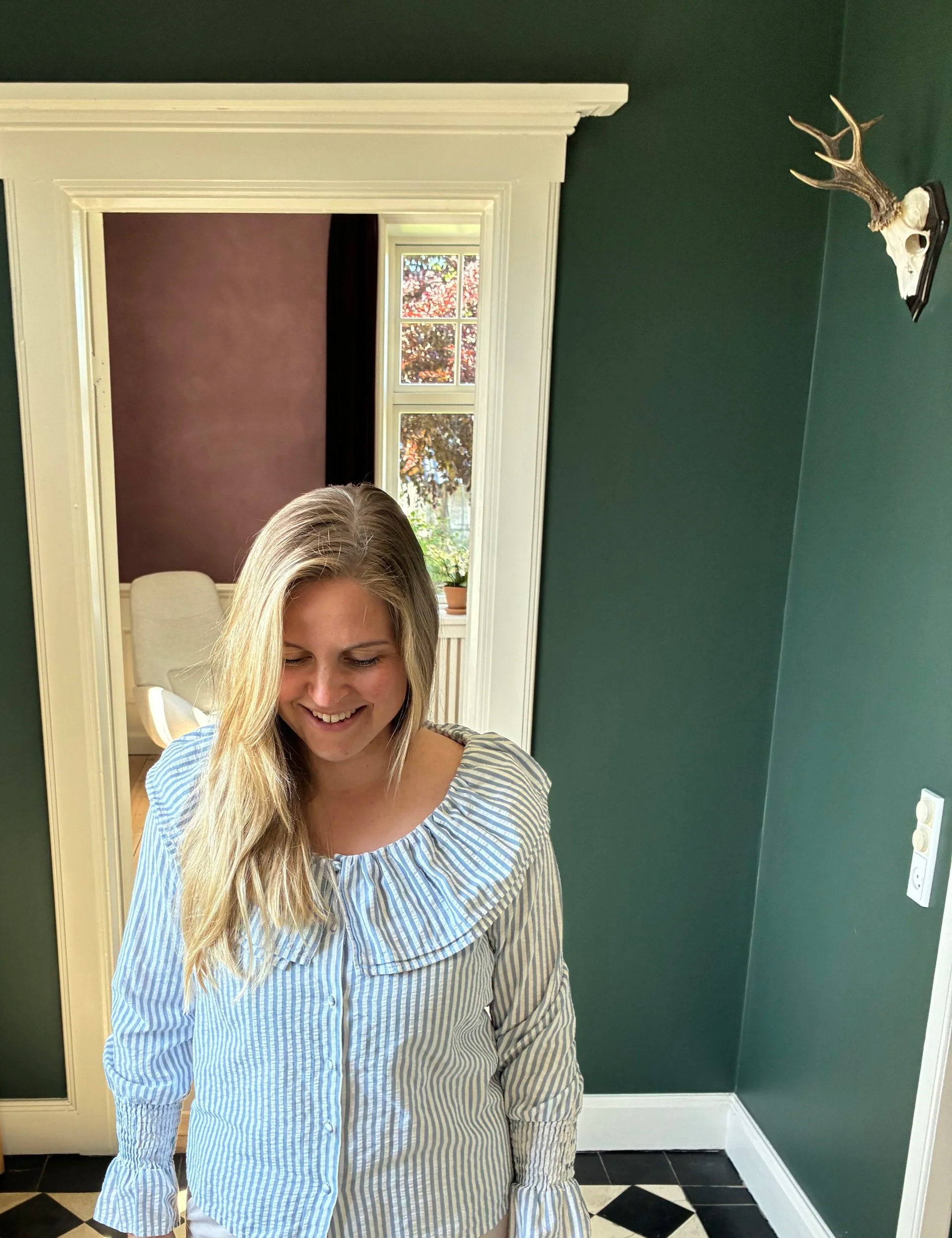 En kvinde med blondt hår, der smiler, står foran et spejl og tager en selfie i et lyst værelse med grøn væg, hvid ramme omkring spejlet, og et vindue med udsigt til farverige løvtræer.