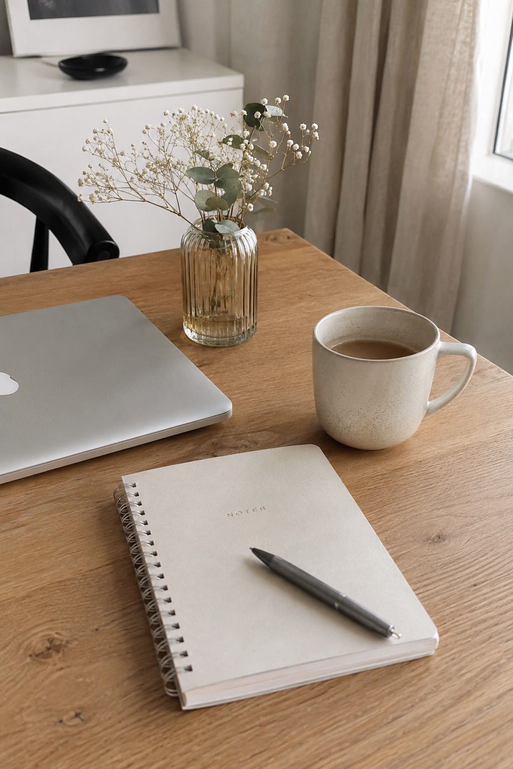Et bord med en notesbog, en kuglepen, en kop kaffe, en buket blomster i en vase, et MacBook og et vindue med gardiner.