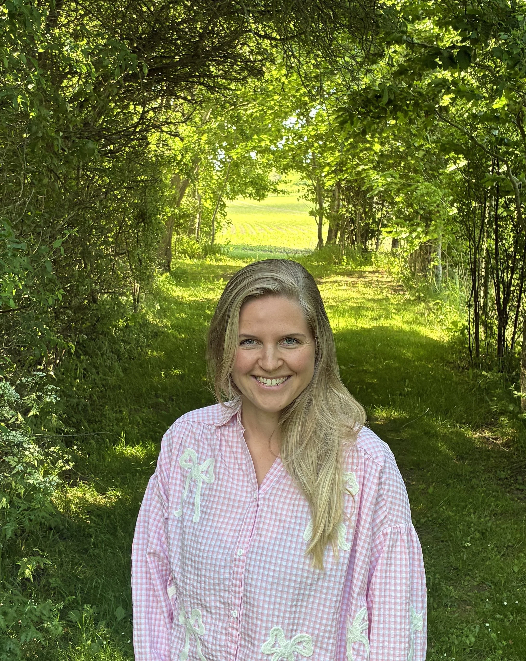 Kvindelig person med lyst hår smiler foran en grøn skov i sollys.